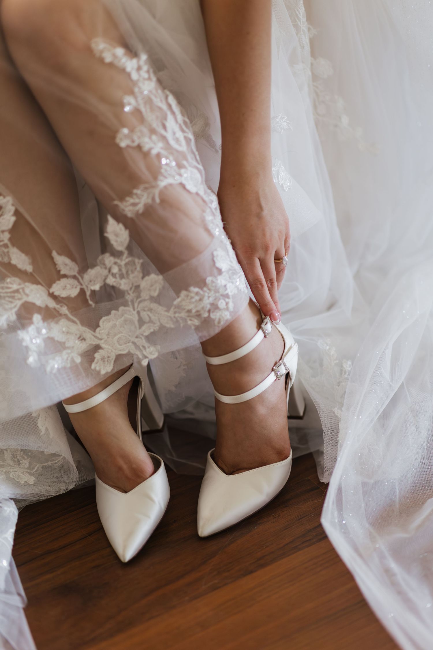 Bride fastening white satin block heel shoes with ankle straps, wearing a lace wedding gown, seated on wooden floor.