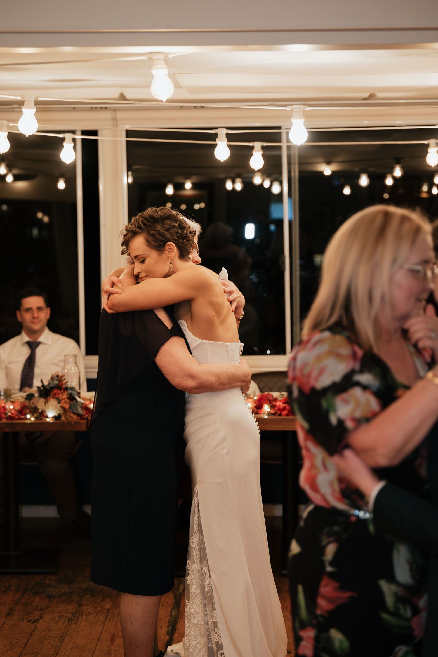 Bride and mother share an emotional first dance hug at wedding reception with warm string lights glowing overhead.