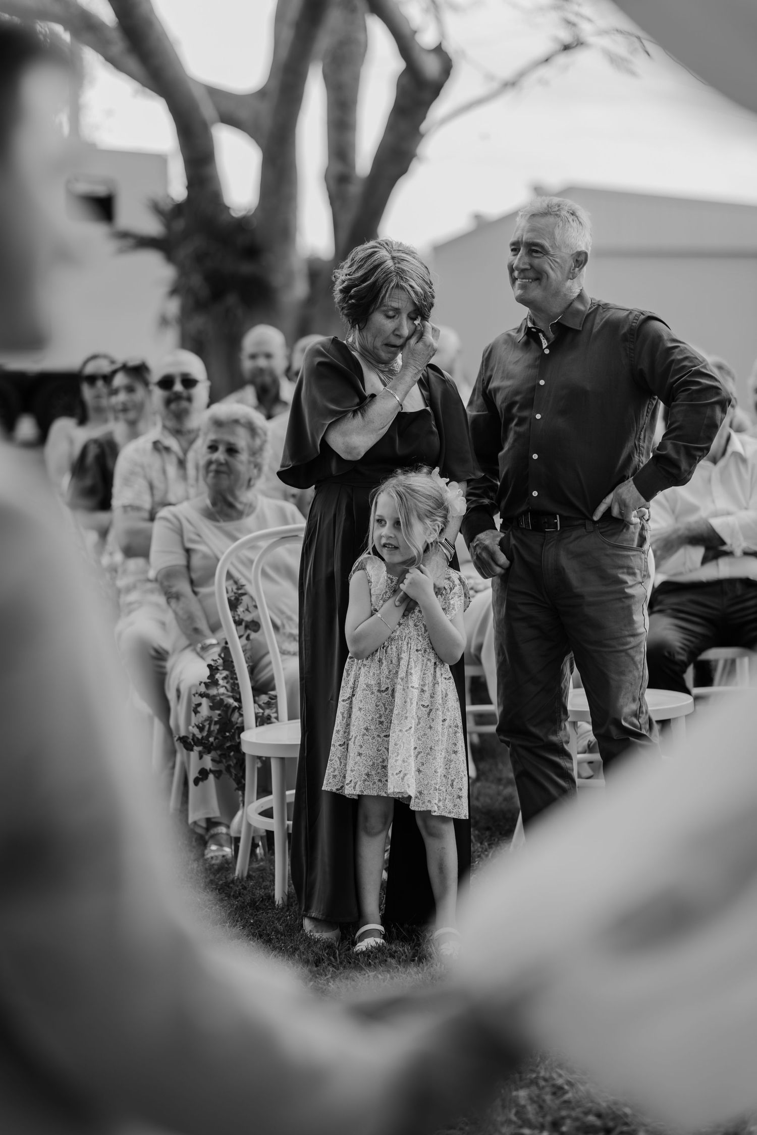 Black and white photo of emotional wedding guests, woman wiping tears beside a young girl clapping at an outdoor ceremony.