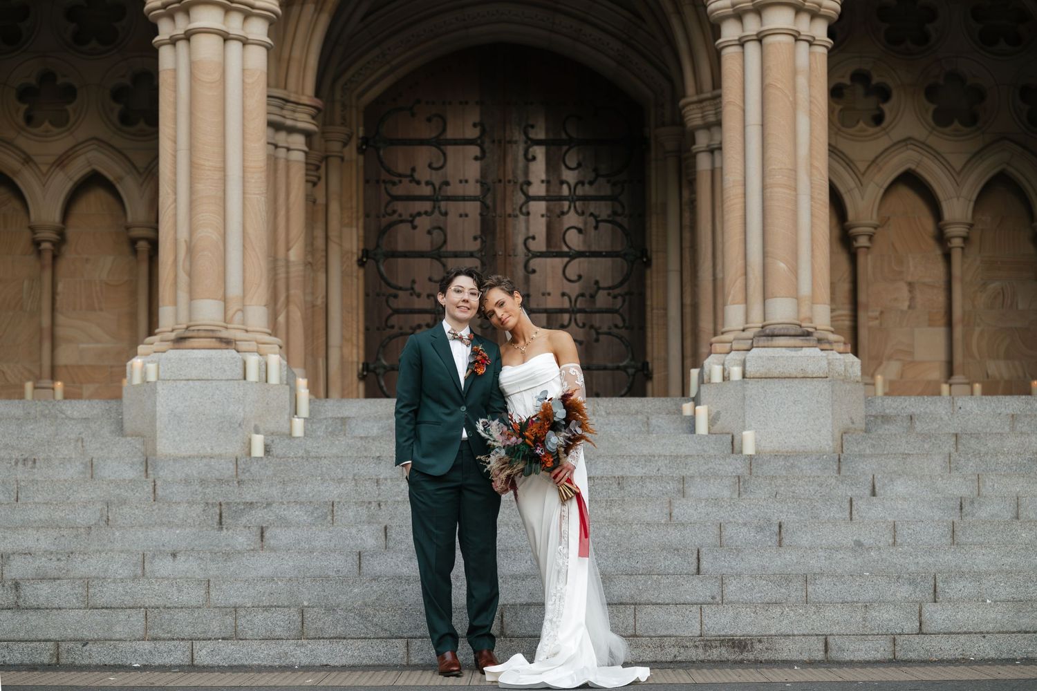Bride and groom posing on stone steps before ornate church doors on their wedding day.