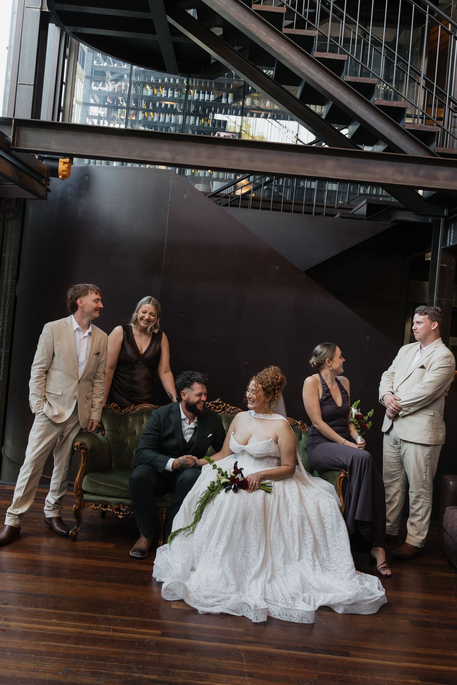 Bride and groom seated on green velvet sofa surrounded by wedding party in industrial venue with spiral staircase.