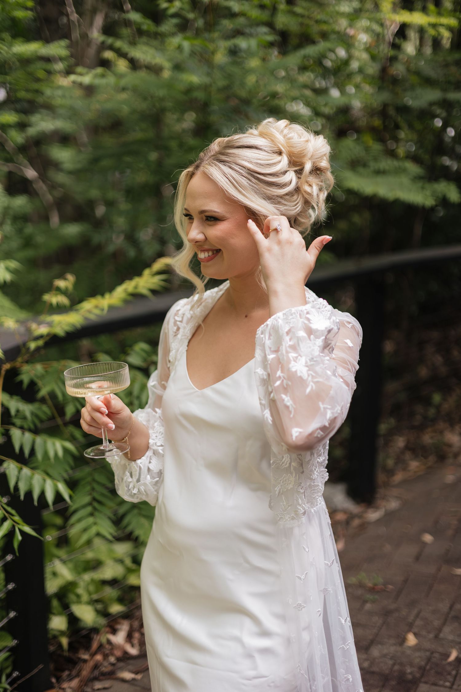 Smiling bride in white lace gown holding champagne coupe glass outdoors among lush green ferns.