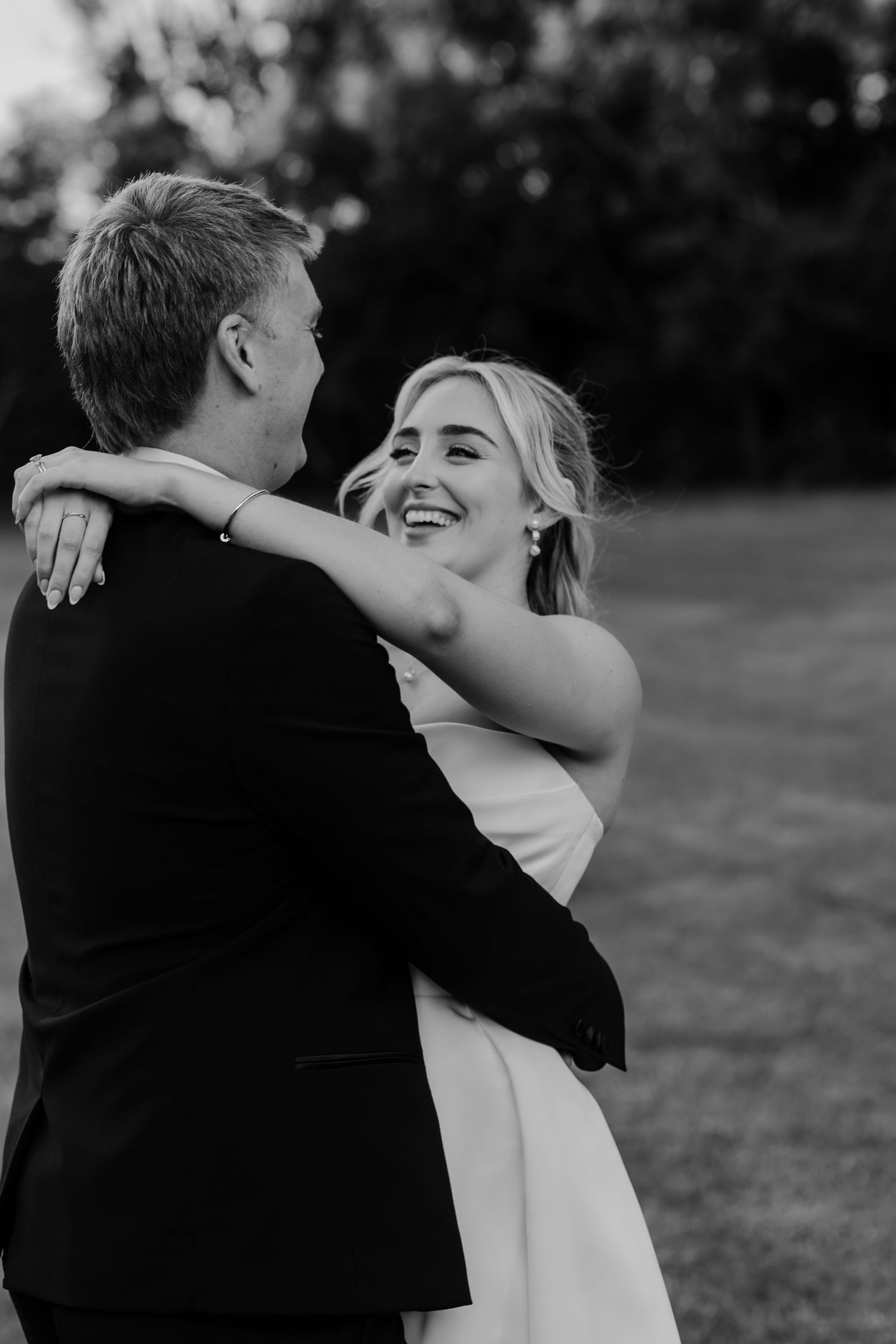 Black and white photo of a smiling bride embracing her groom outdoors during a romantic wedding dance.