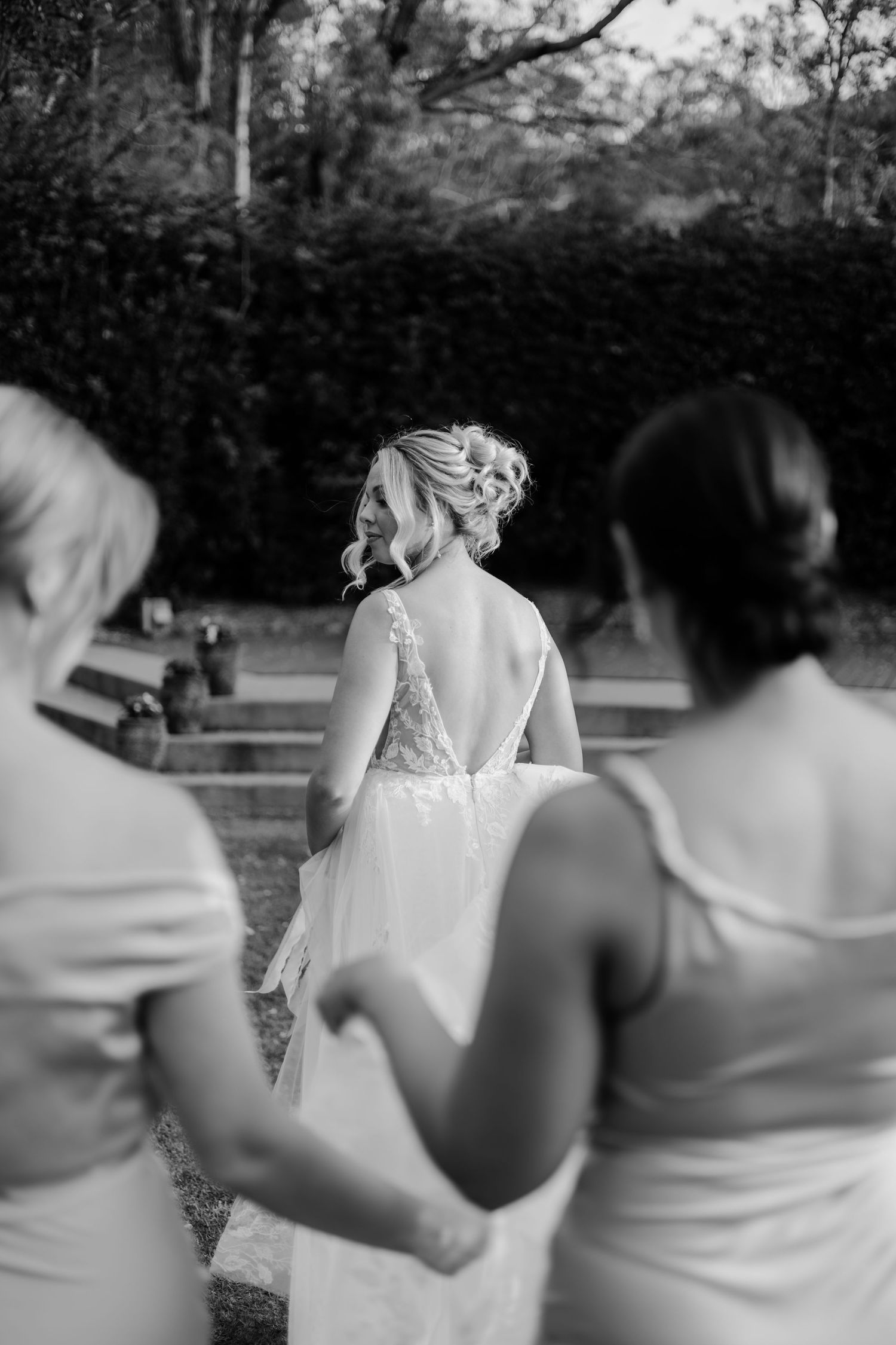 Black and white photo of a bride in a backless lace gown walking with two bridesmaids in a garden.
