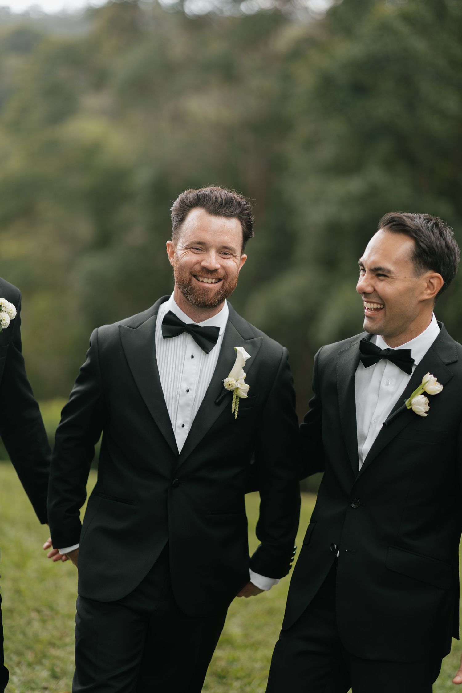Two groomsmen in black tuxedos with bow ties and white boutonnieres smile and laugh outdoors at a wedding.