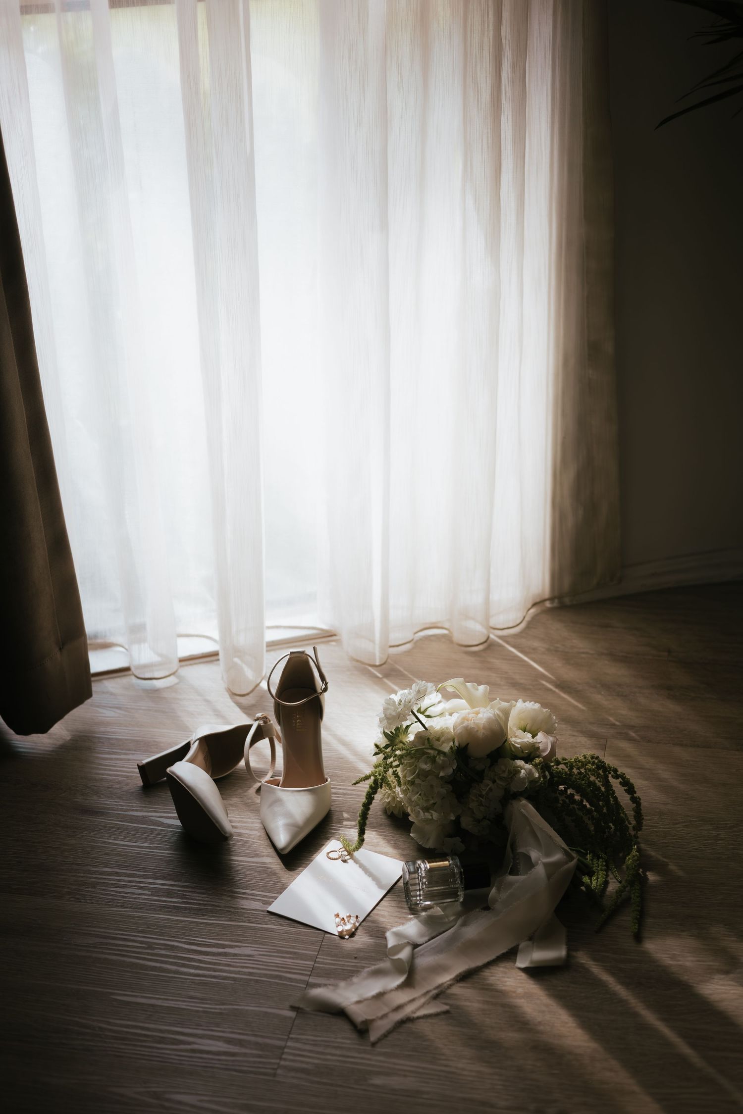 Bridal flat lay with white flower bouquet, heels, sunglasses, and rings on wooden floor by sheer curtains.