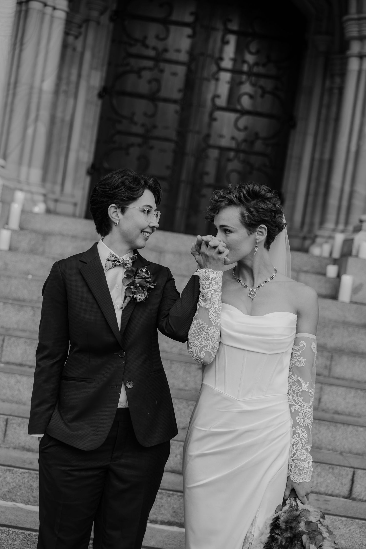 Black and white photo of two brides in wedding attire smiling at each other on church steps.