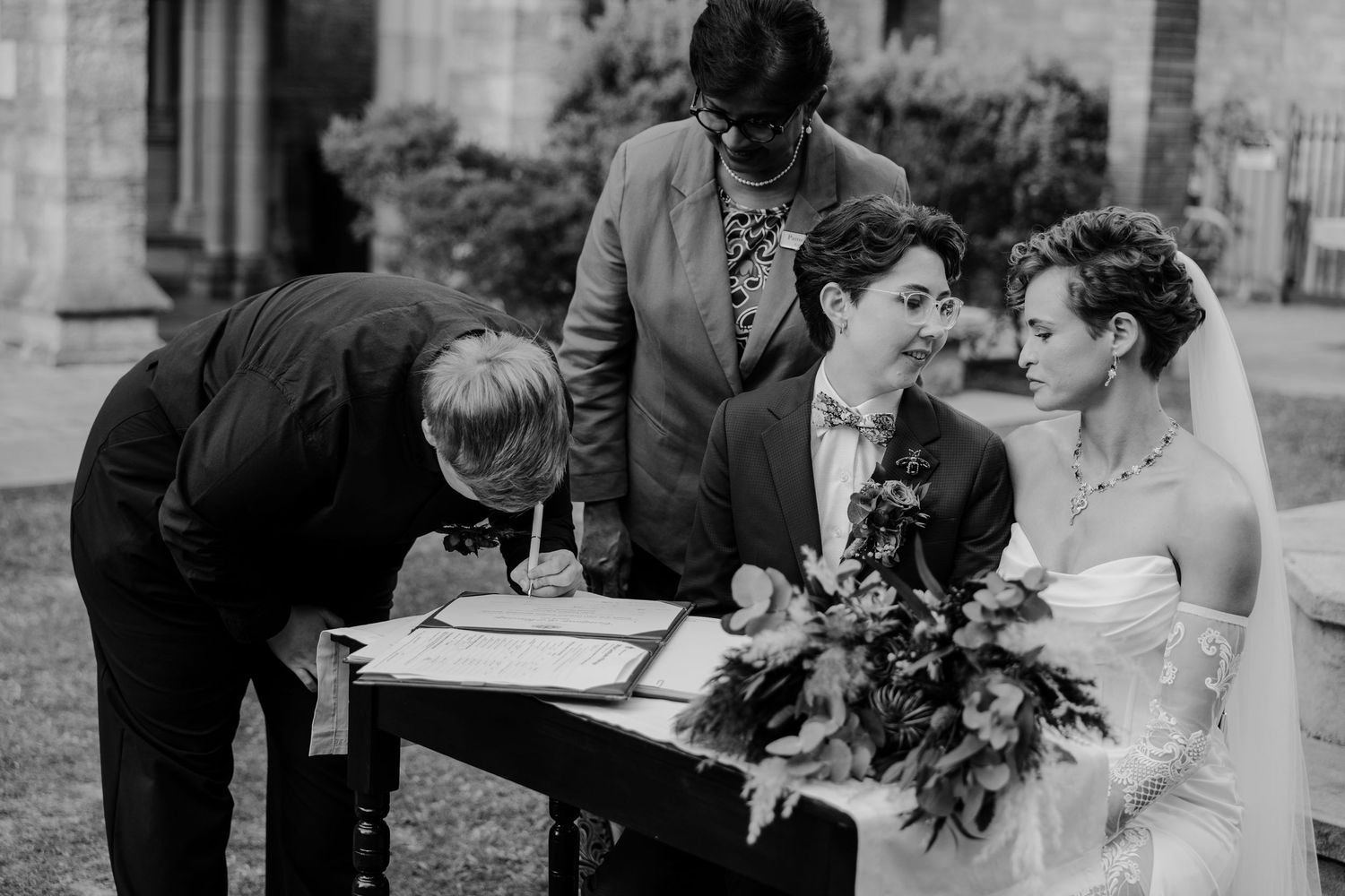 Black and white wedding photo of couple signing marriage register with officiant and guest outdoors.