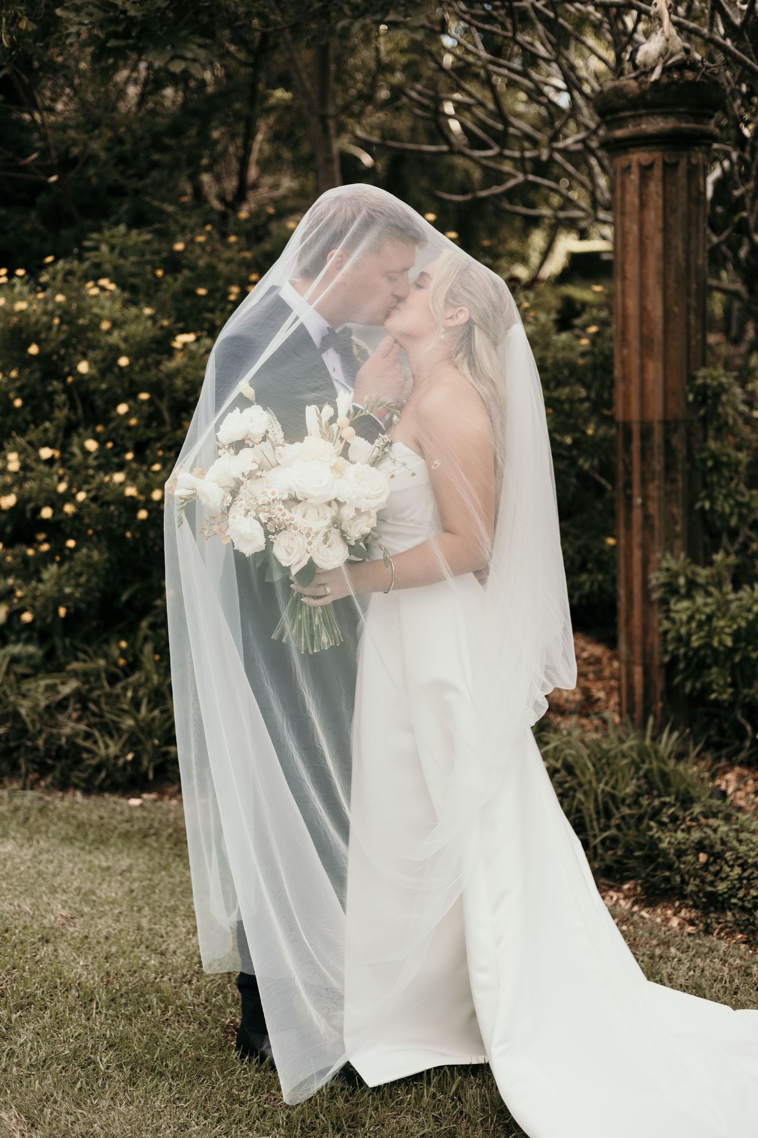 Bride and groom kissing under a flowing white veil, bride holding white floral bouquet in a lush garden setting.