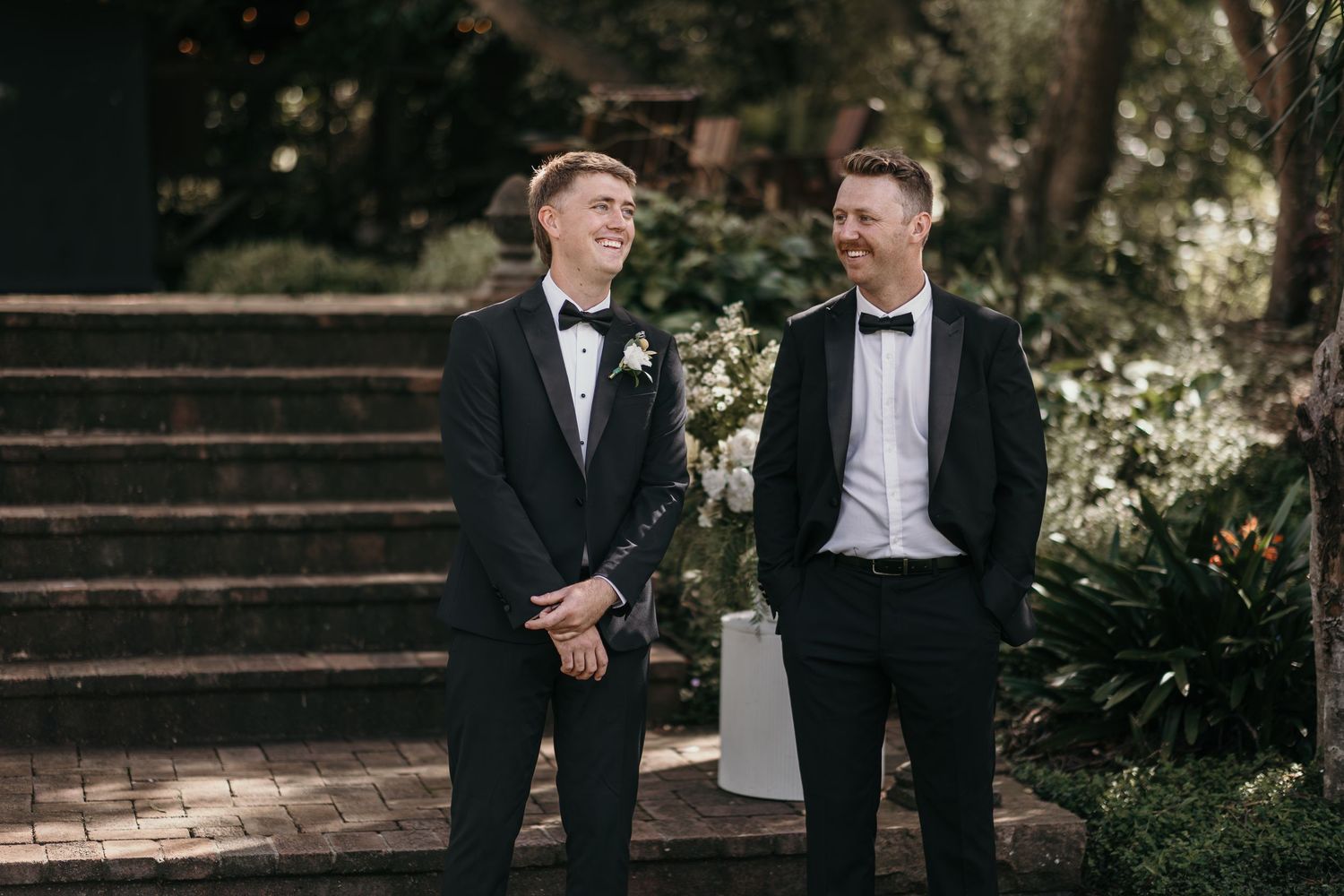 Two grooms in black tuxedos and bow ties smiling at each other outdoors on their wedding day.