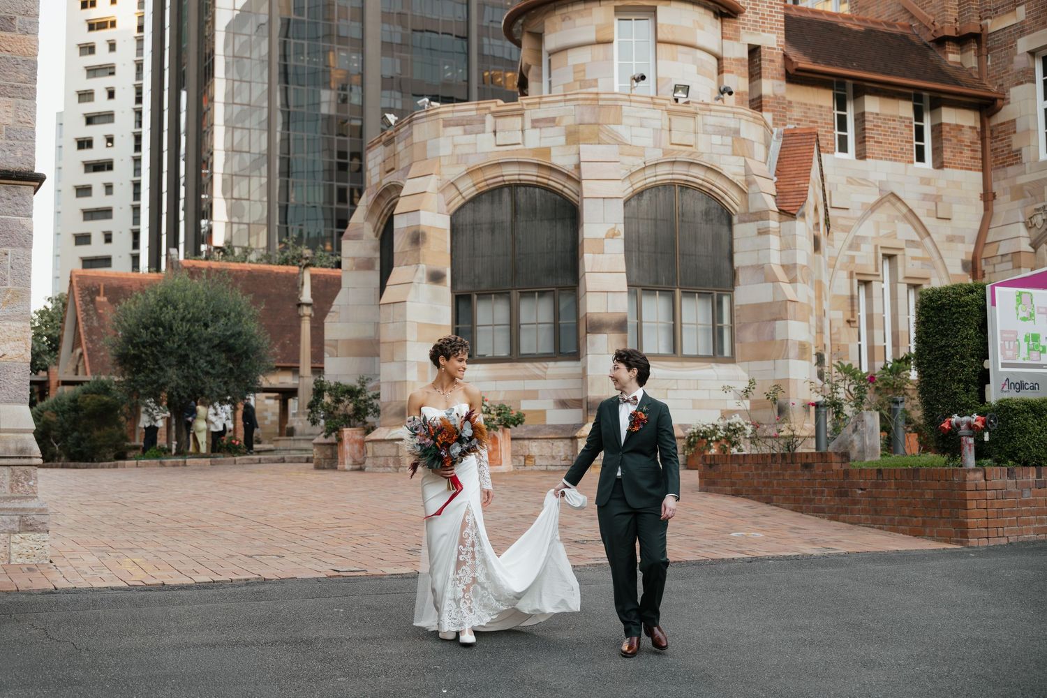 Bride and groom walk hand-in-hand outside a historic sandstone church with city skyscrapers in the background.
