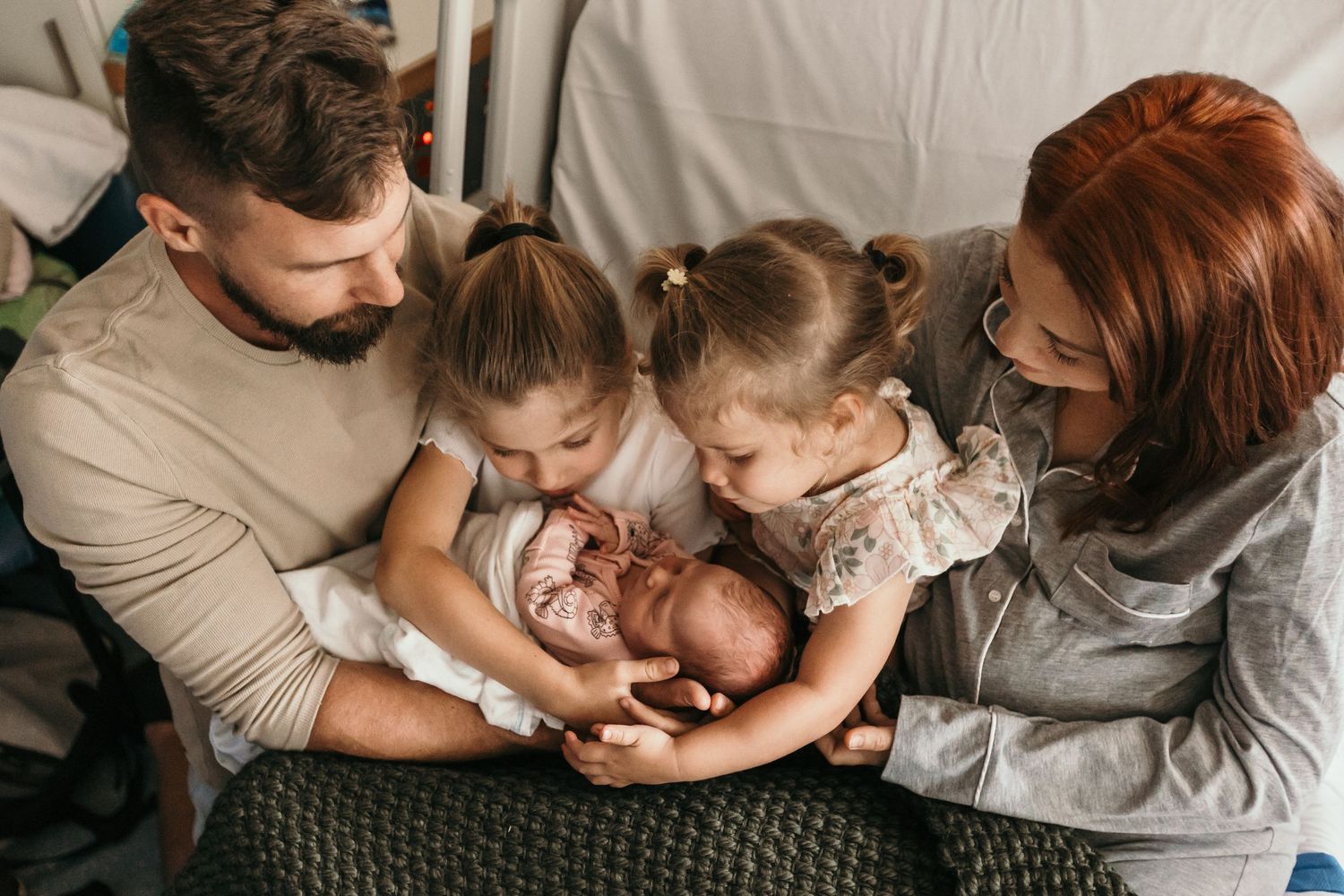 A loving family of four admires their newborn baby together, with two young sisters gently cradling the infant.