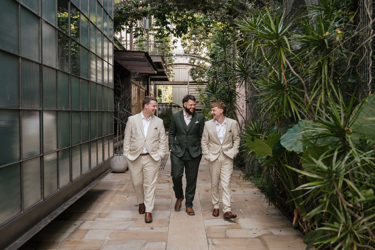 Three men in suits walking confidently down a lush garden pathway flanked by tropical plants and glass walls.