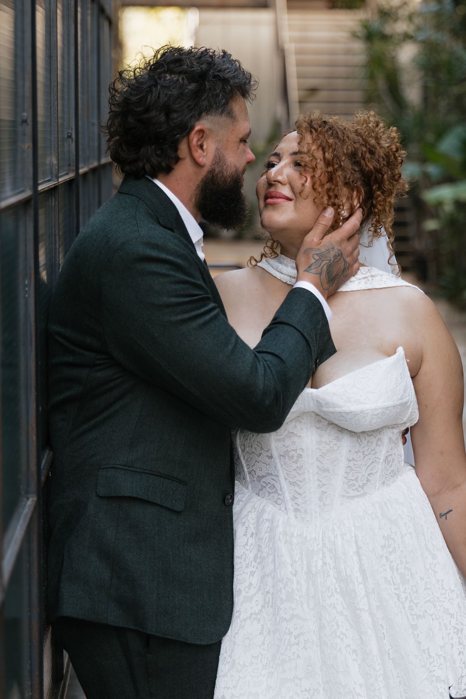 Bearded groom in dark suit tenderly holds brides face as she smiles in white lace wedding dress outdoors.