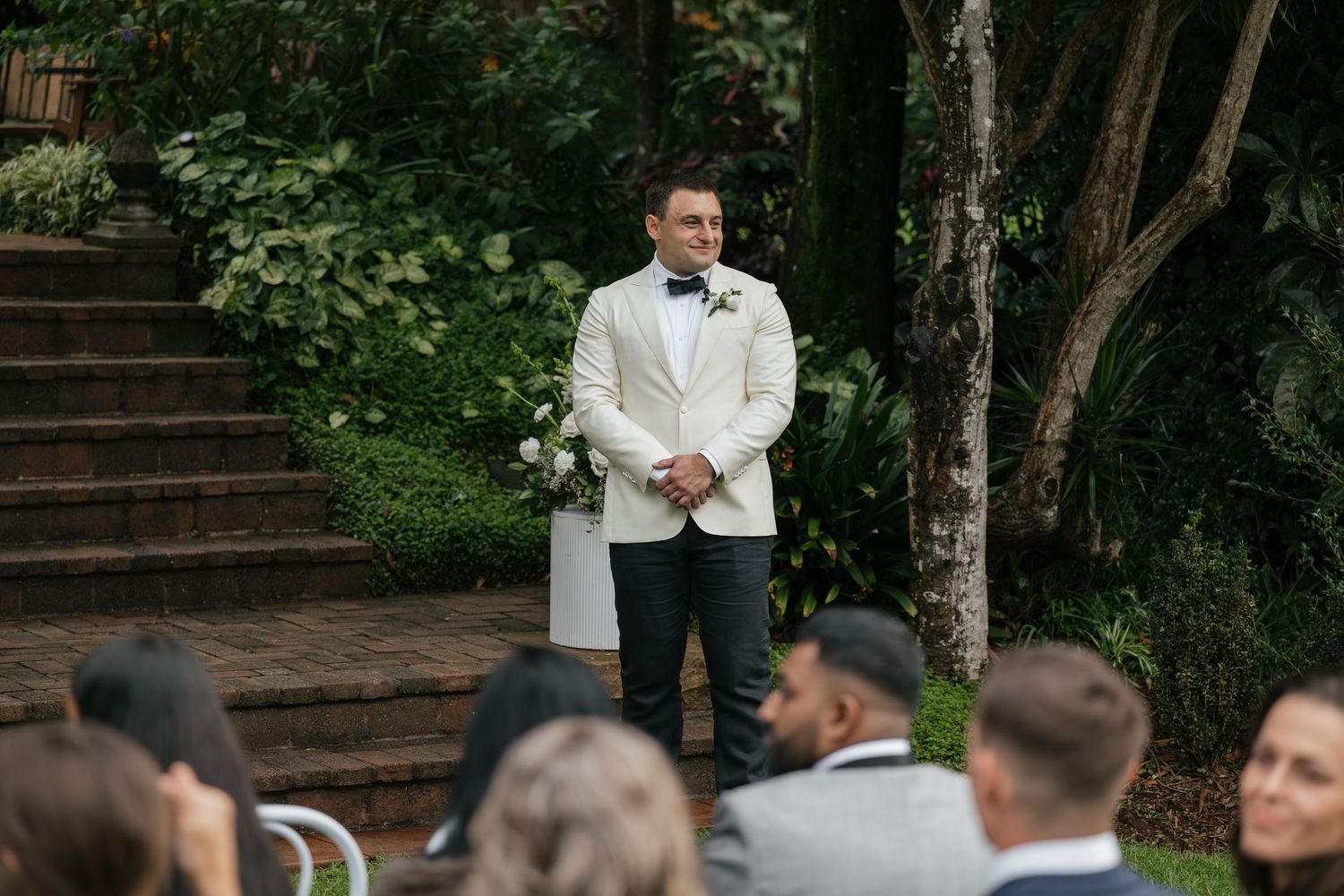 Groom in white tuxedo jacket stands at outdoor wedding ceremony surrounded by lush greenery, awaiting his bride.