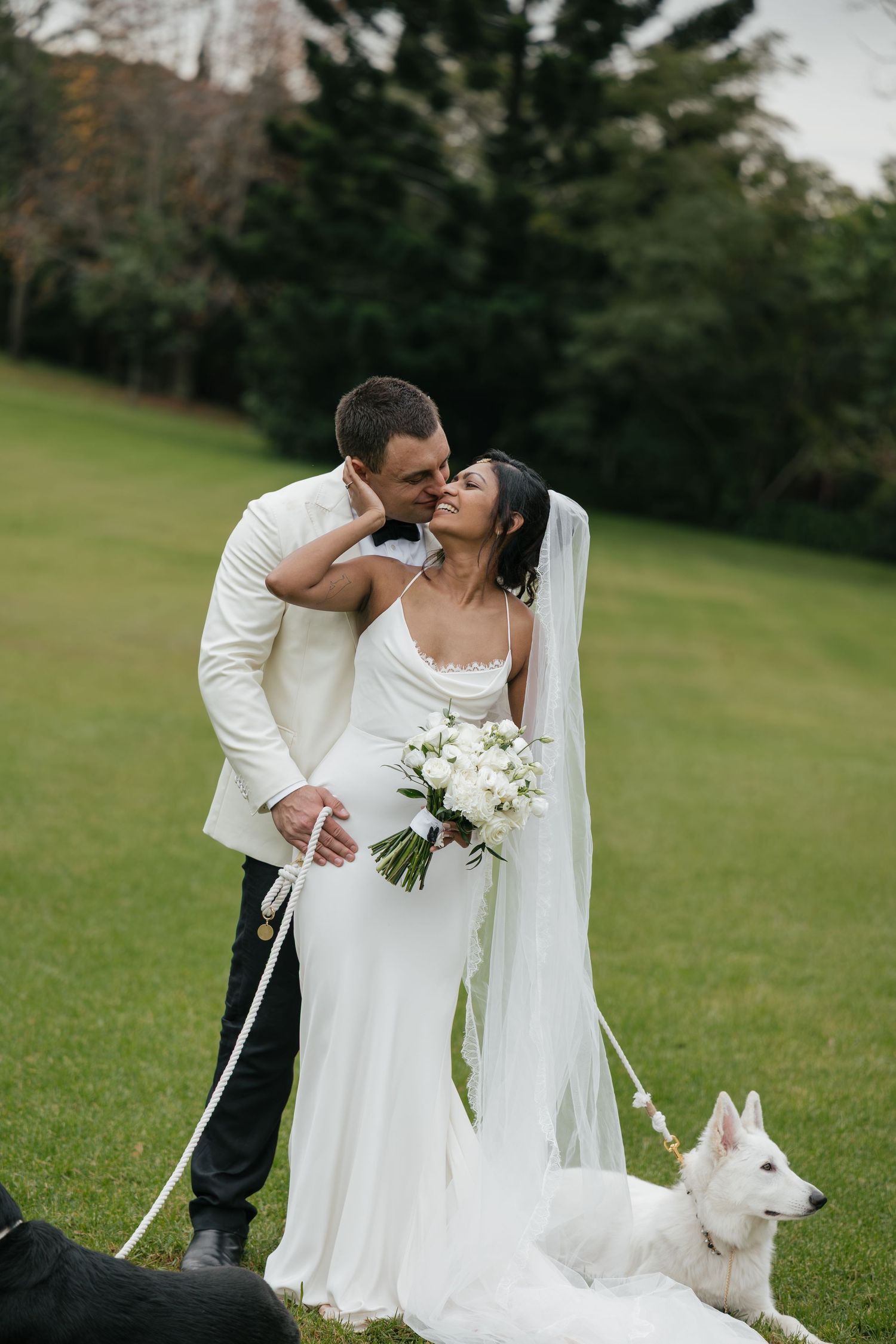 Bride and groom share a kiss on green lawn, bride holds white bouquet, white dog sits nearby.