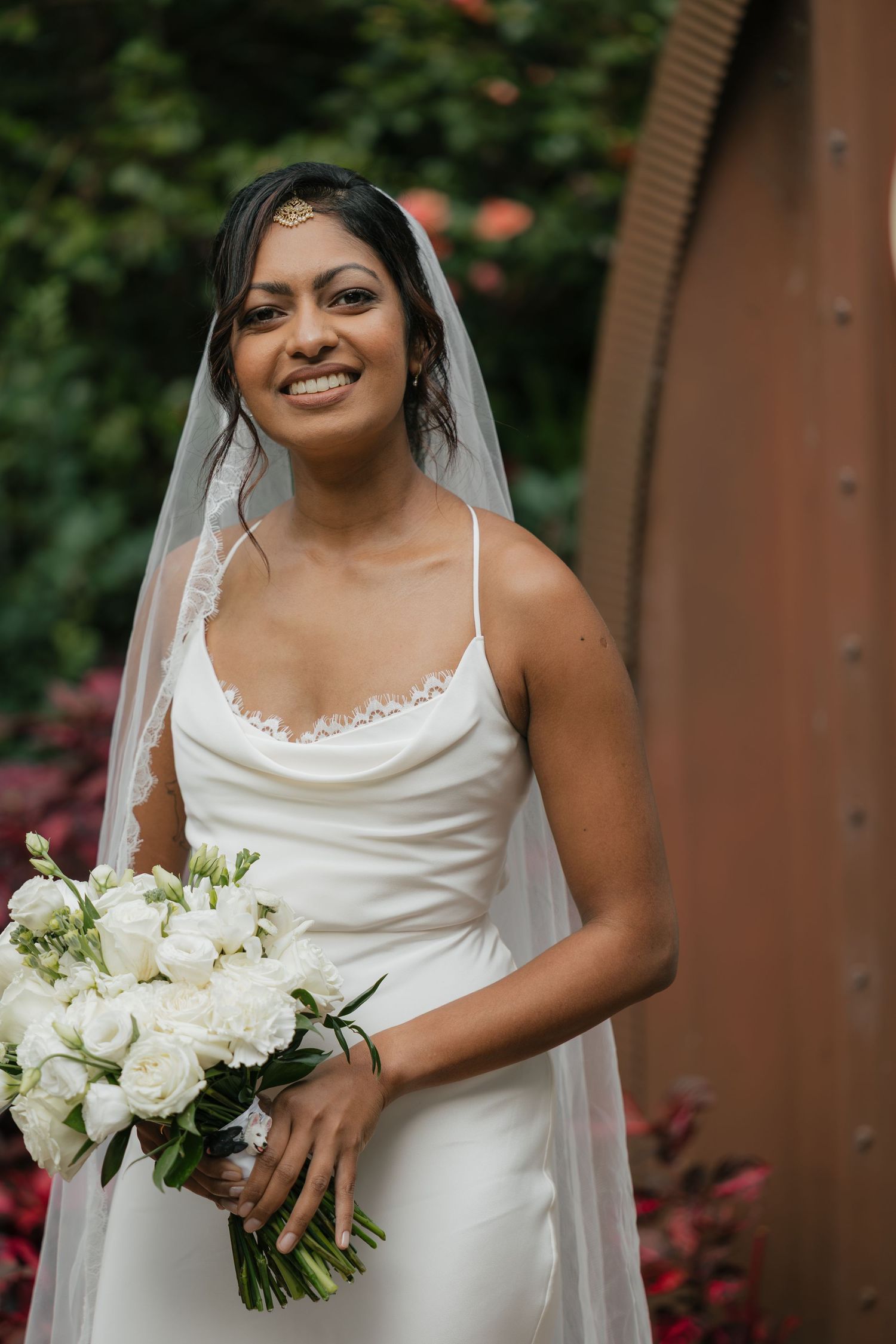 Smiling bride in white cowl-neck gown and veil holds white rose bouquet outdoors amid lush greenery.