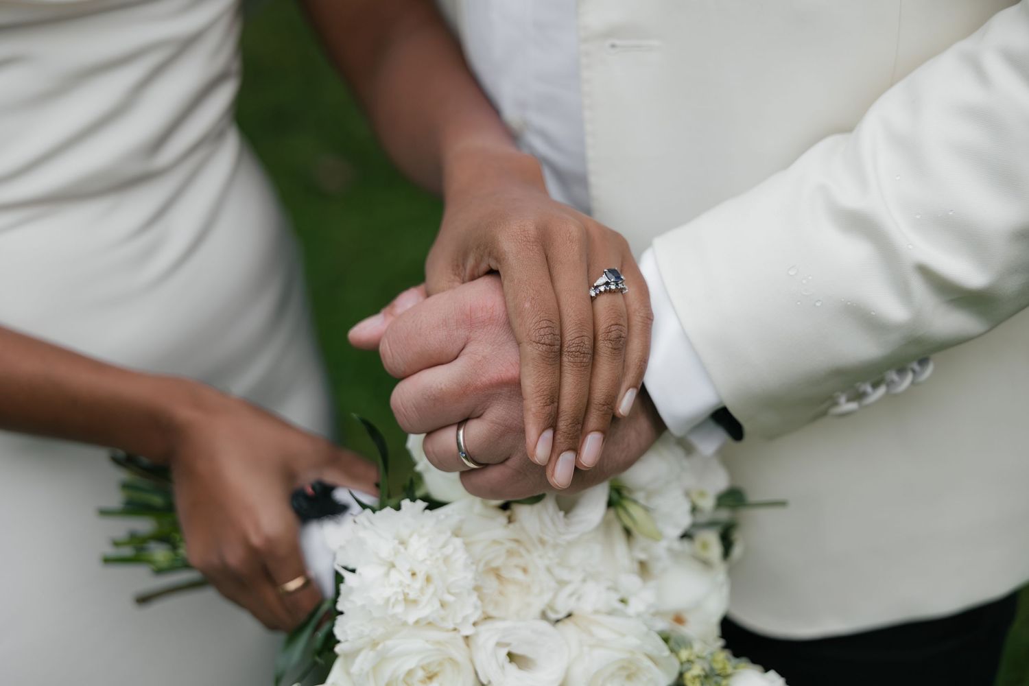 Bride and groom holding hands showing wedding rings with white floral bouquet at wedding ceremony.