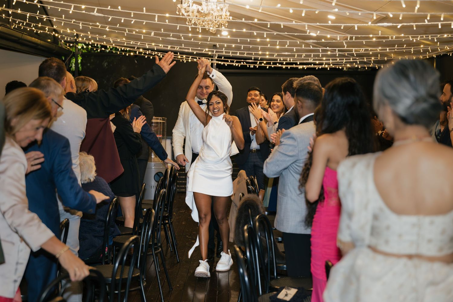 Bride in white mini dress dancing joyfully with guests at a fairy light-adorned wedding reception venue.