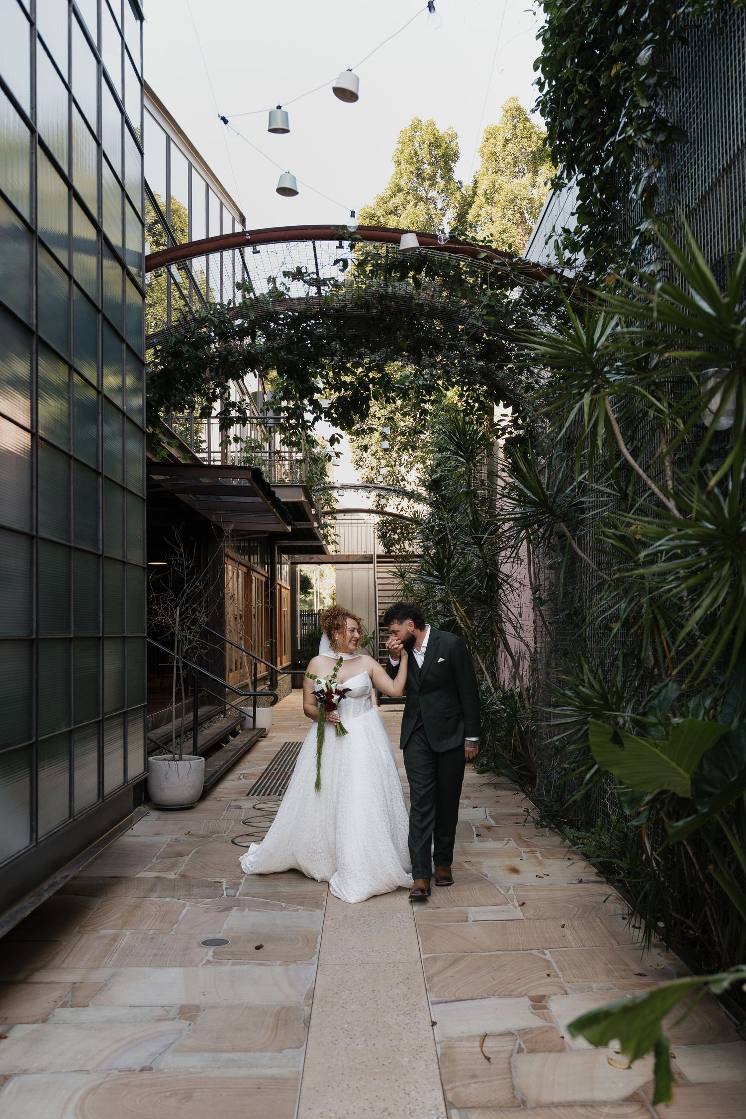 Bride and groom walk together under lush green archway in elegant outdoor courtyard during wedding celebration.