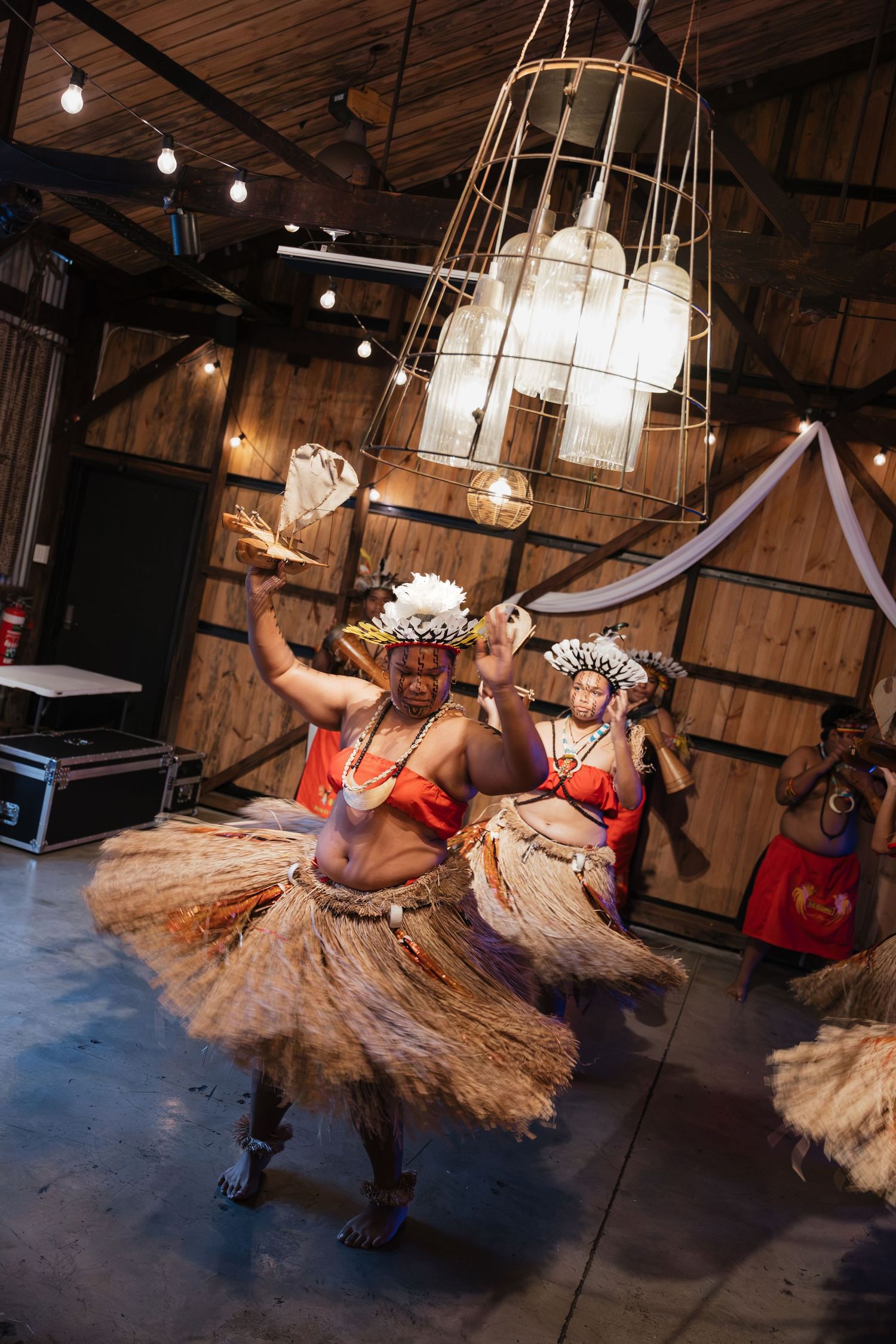 Traditional Polynesian dancers in grass skirts and floral headdresses perform energetically indoors under warm pendant lights.