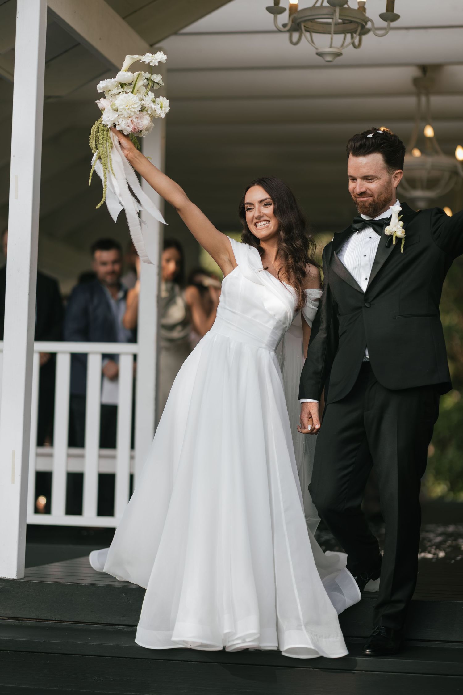 Bride raises bouquet triumphantly as groom in black tuxedo smiles beside her on wedding venue steps.