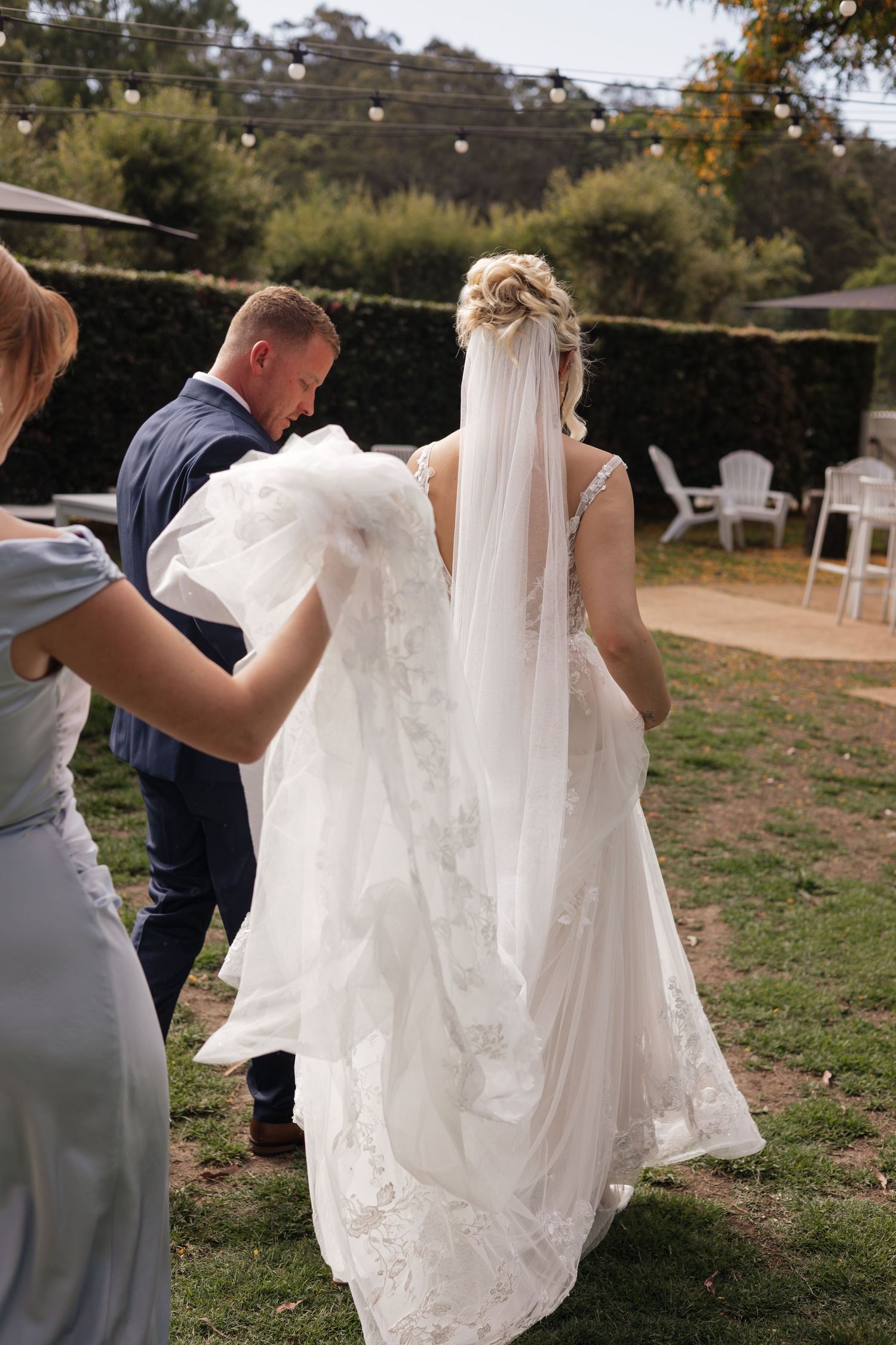 Bride in lace wedding gown with veil walks outdoors, assisted by bridesmaid and groomsman in garden setting.