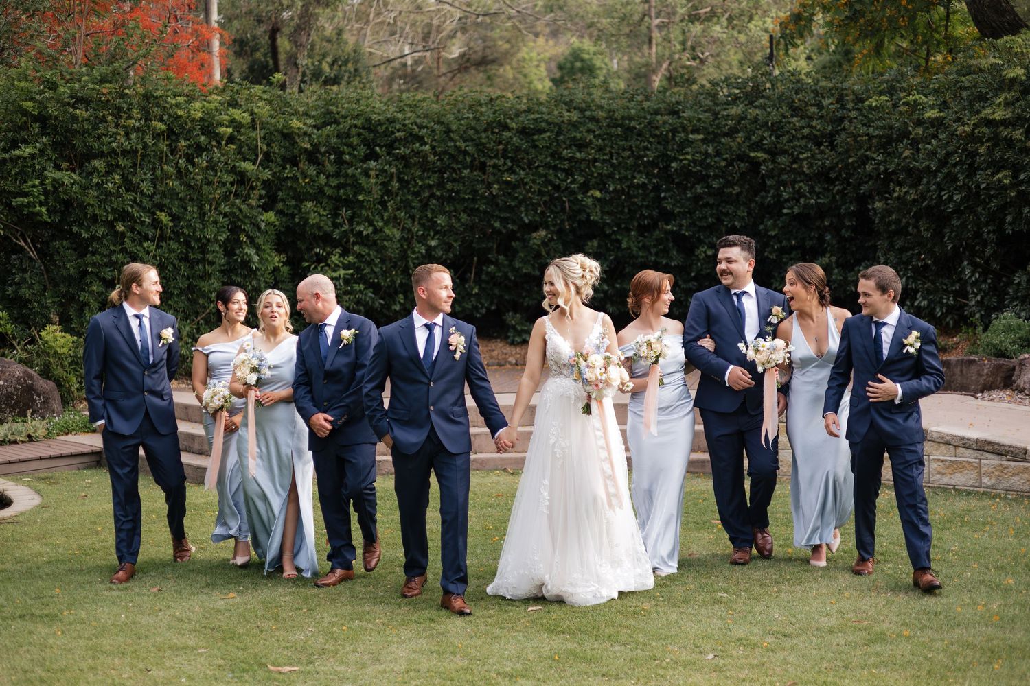 Wedding party walks on green lawn, bride in white gown with bridesmaids in blue and groomsmen in navy suits.