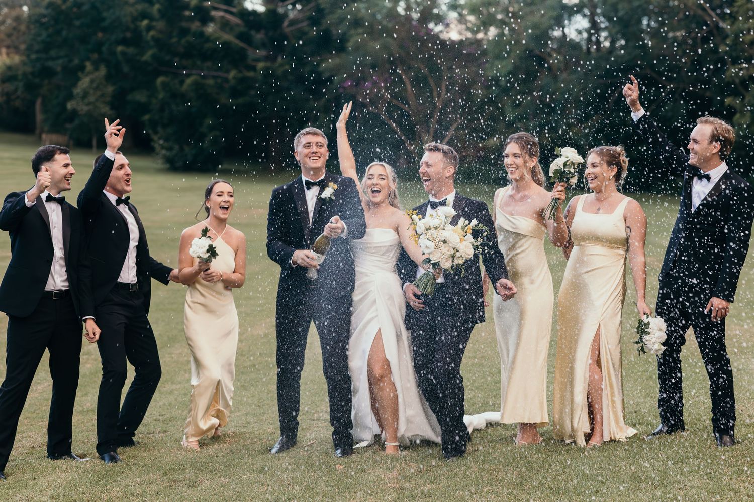 Wedding party celebrating outdoors on grass, bride and groom with bridesmaids and groomsmen in gold and black, confetti flying.