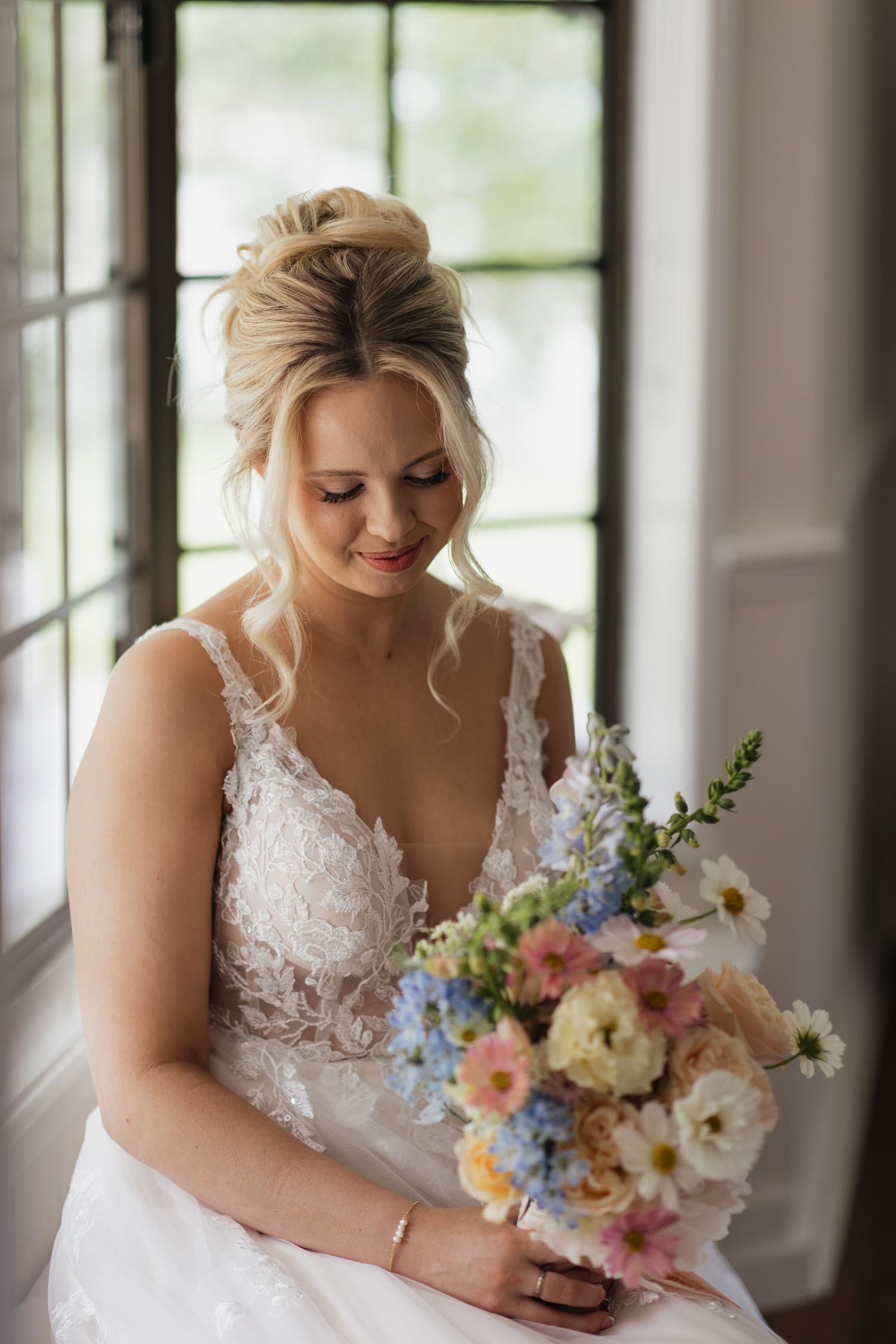Bride in lace wedding gown holding colorful spring bouquet with blue, pink, and white flowers, smiling softly indoors.