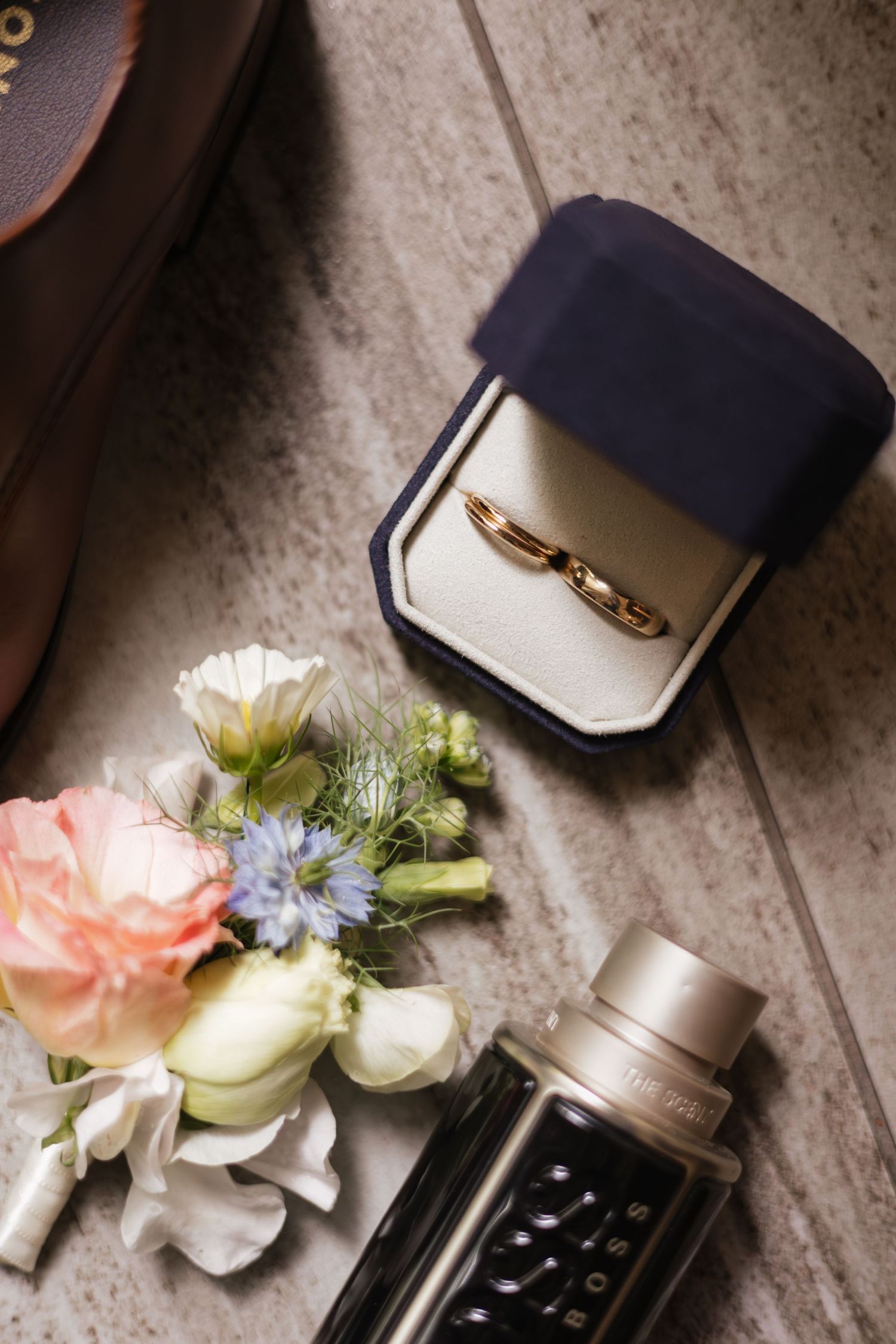 Gold wedding rings in a navy blue velvet box beside a floral boutonniere and cologne bottle on a wooden surface.