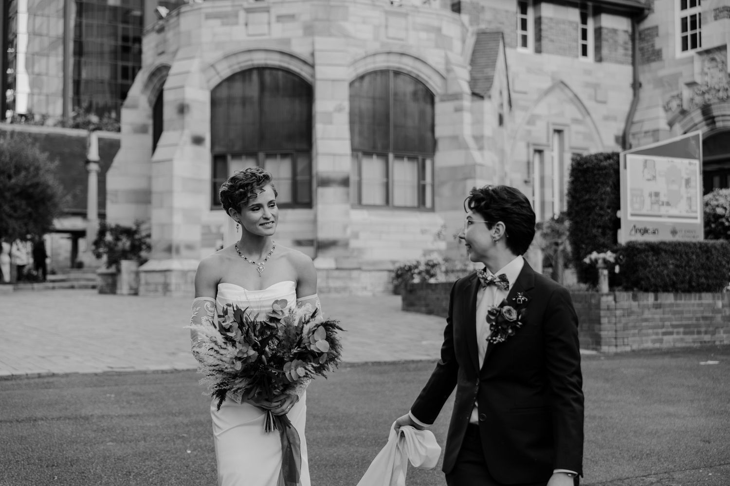 Black and white photo of a bride holding a bouquet and groom in a suit smiling at each other outside a stone building.