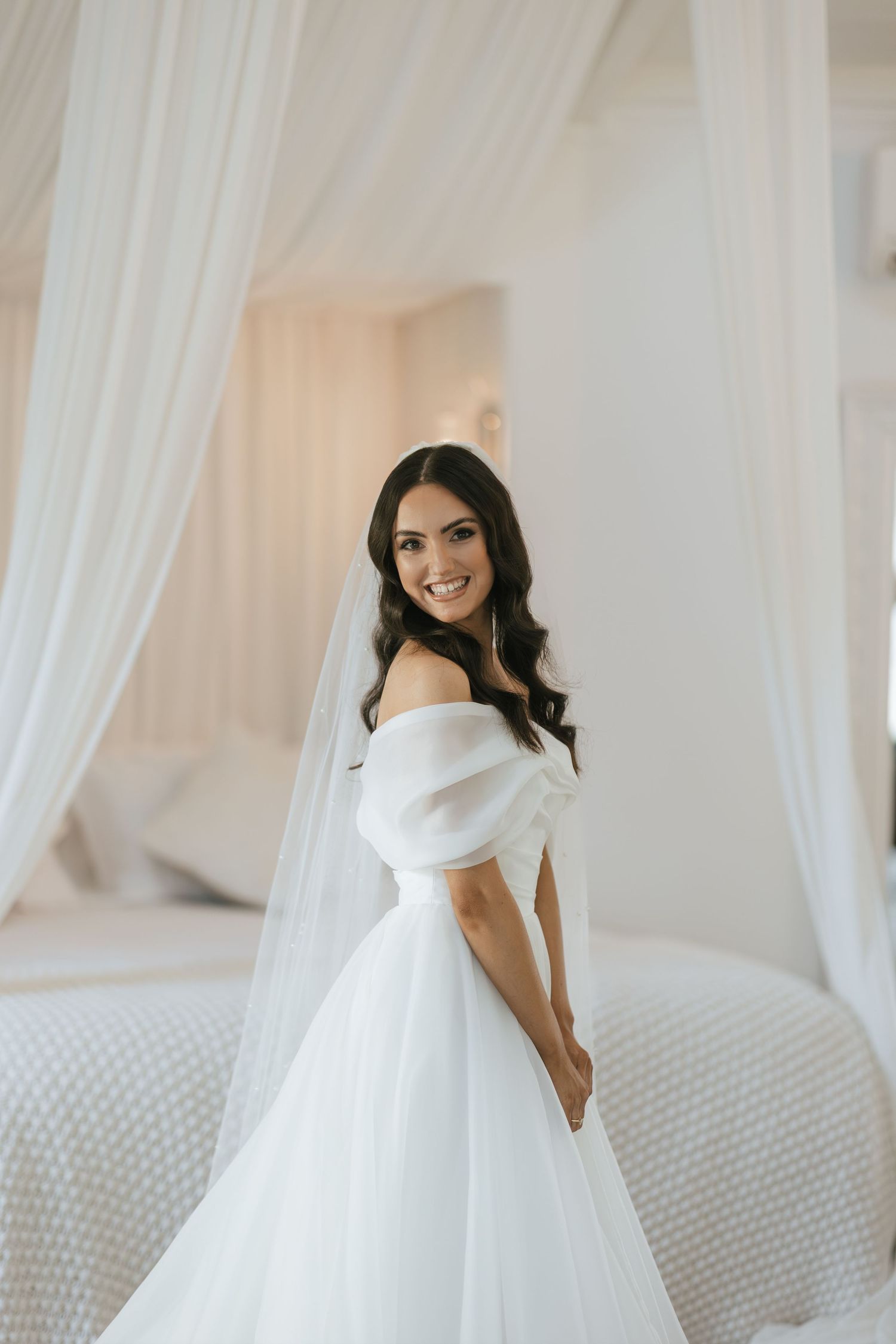 Smiling bride in elegant off-shoulder white ball gown with veil, posing in a bright, airy bridal suite.
