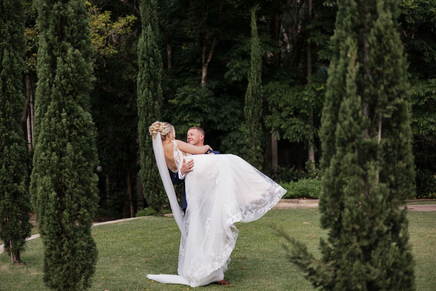 Groom lifting bride in white wedding gown among tall cypress trees on lush green lawn at outdoor wedding.