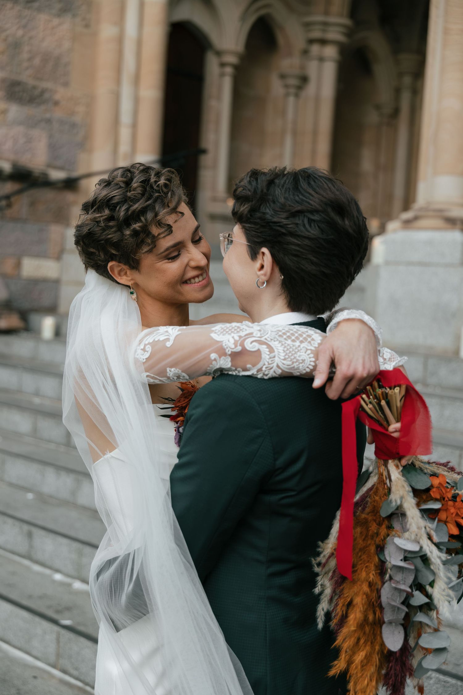 Two brides in wedding attire embrace on stone steps, one in white lace gown with veil, the other in dark suit holding bouquet.