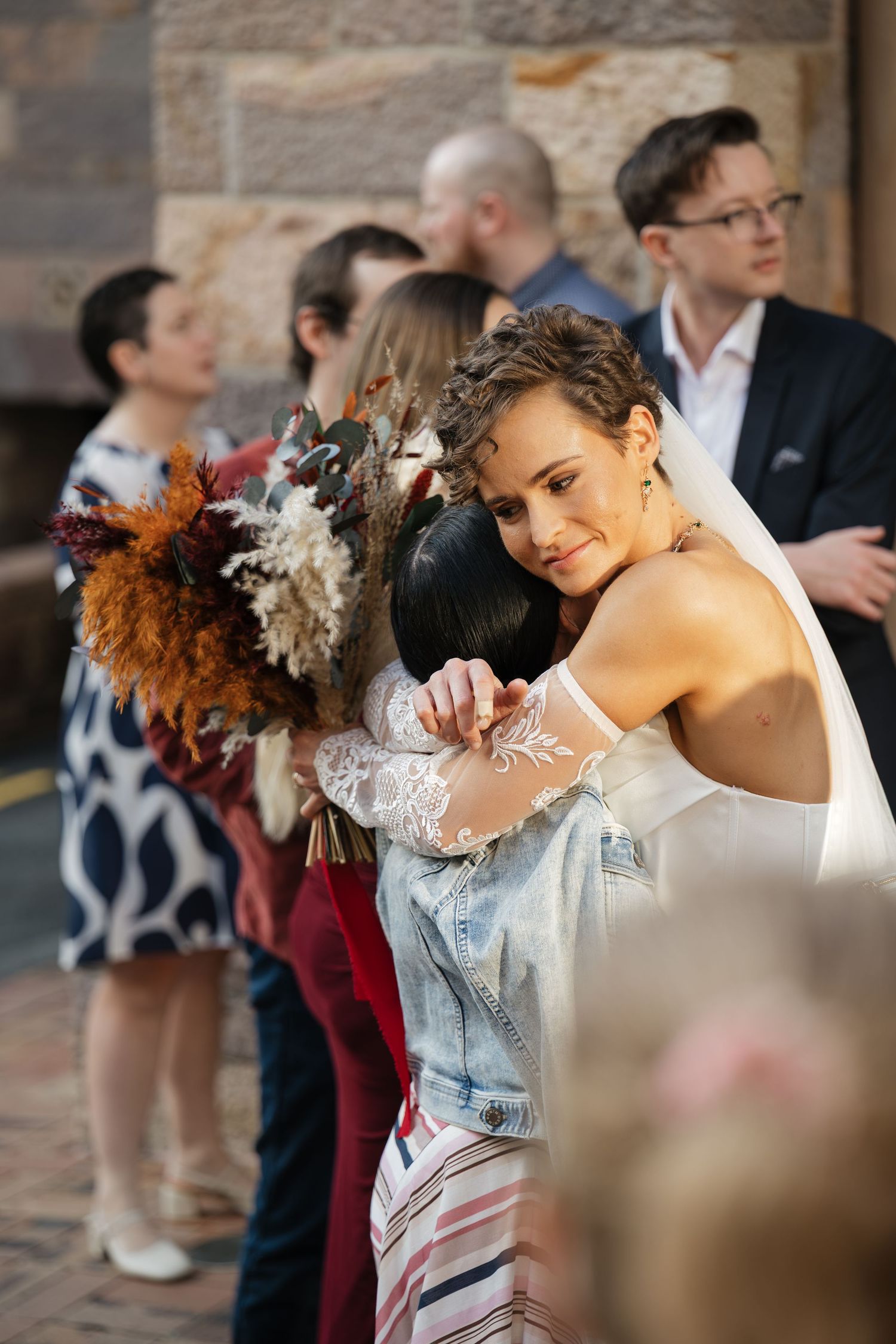 Bride in white dress and veil hugging a guest while holding a dried floral bouquet at an outdoor wedding.