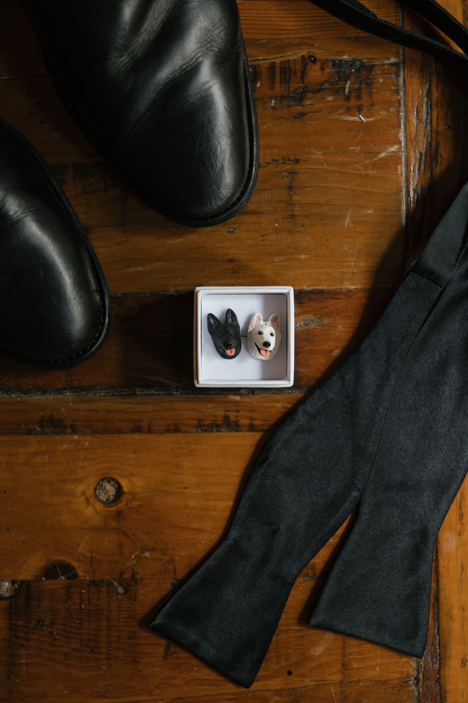 Black leather dress shoes, dog cufflinks in a box, and a charcoal bow tie arranged on a rustic wooden surface.