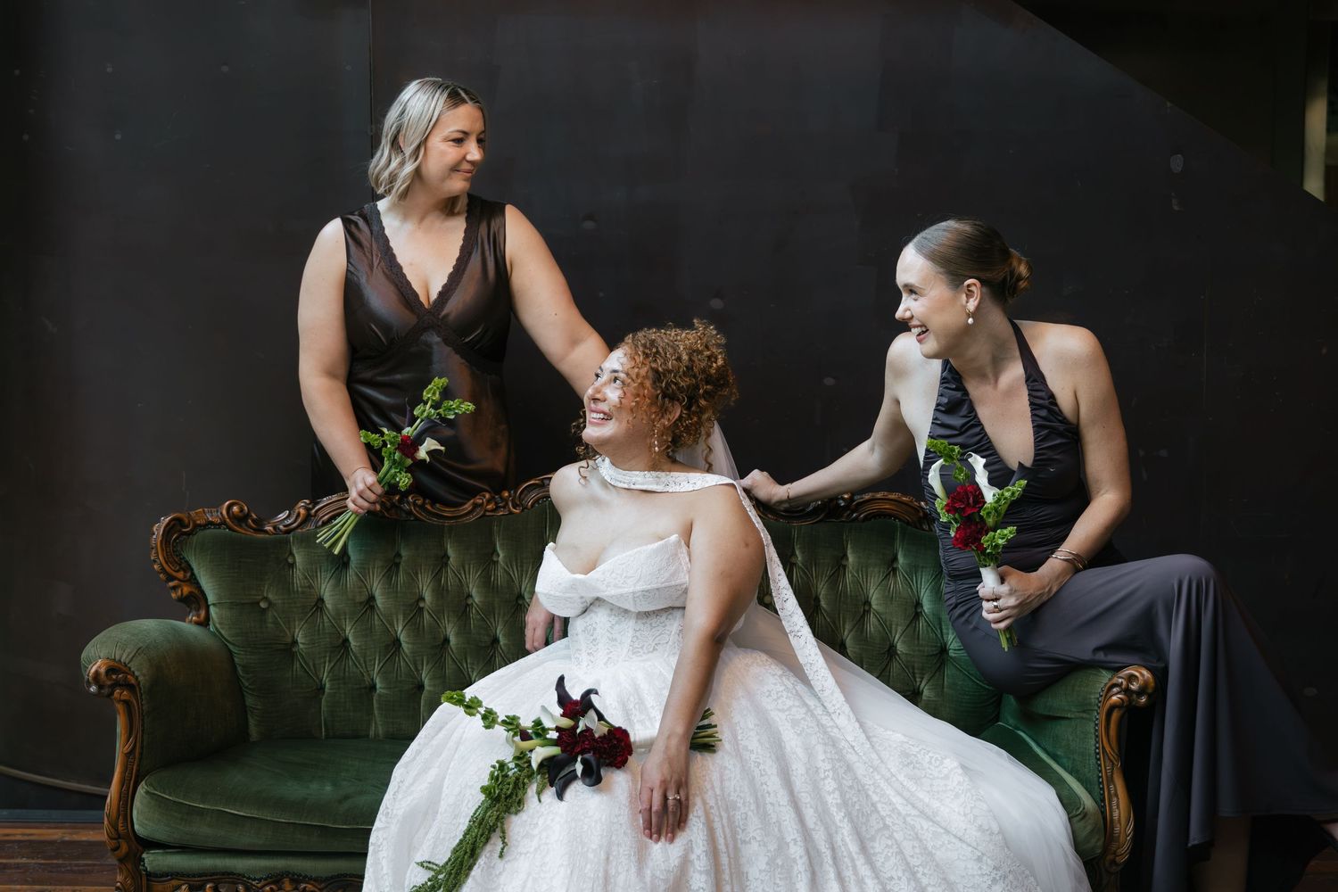 Bride in white gown sits on green velvet sofa with two bridesmaids in gray dresses, laughing together.