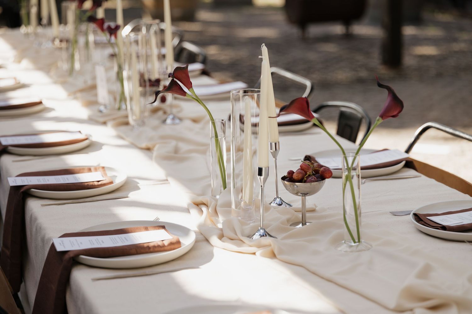 Elegant outdoor dining table set with white linens, wooden chargers, calla lily centerpieces, and candles in natural light.