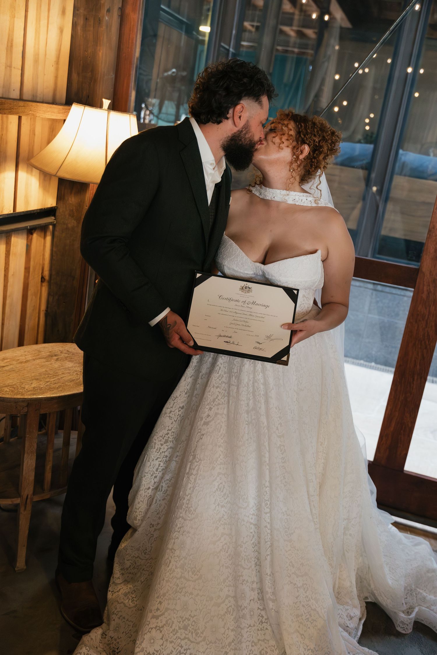 Newlywed couple kissing while holding marriage certificate, bride in lace gown and groom in black suit, rustic indoor venue.