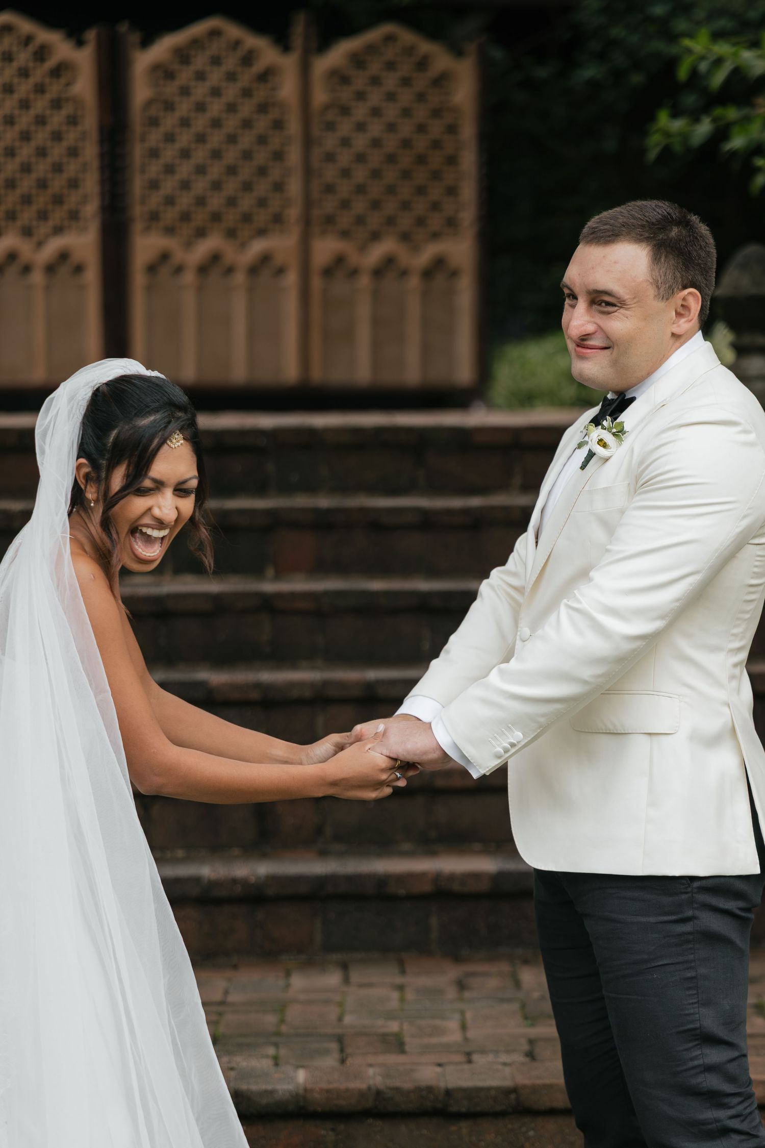 Bride in white gown and veil laughs joyfully holding hands with groom in white tuxedo jacket on brick steps.