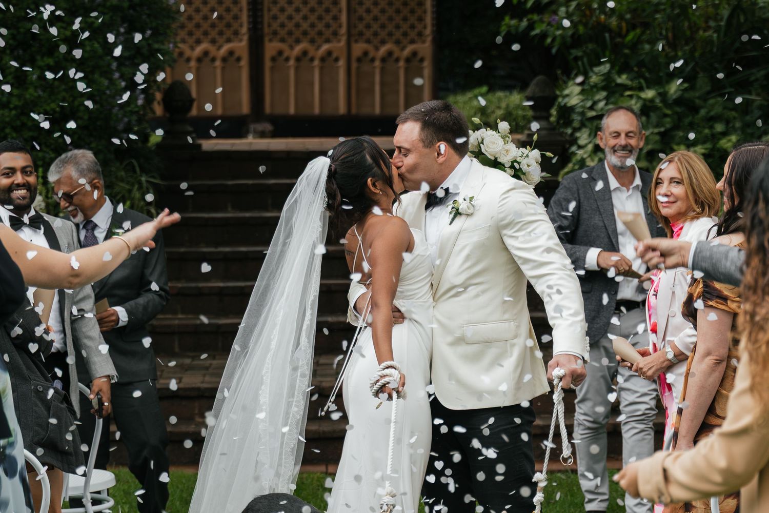 Bride and groom share a kiss while guests celebrate with white confetti at an outdoor wedding ceremony.