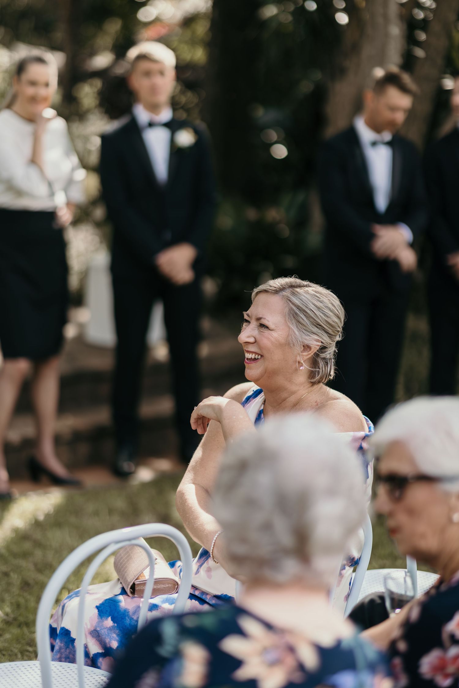 A woman in a floral dress laughs joyfully at an outdoor wedding ceremony with groomsmen standing in the background.