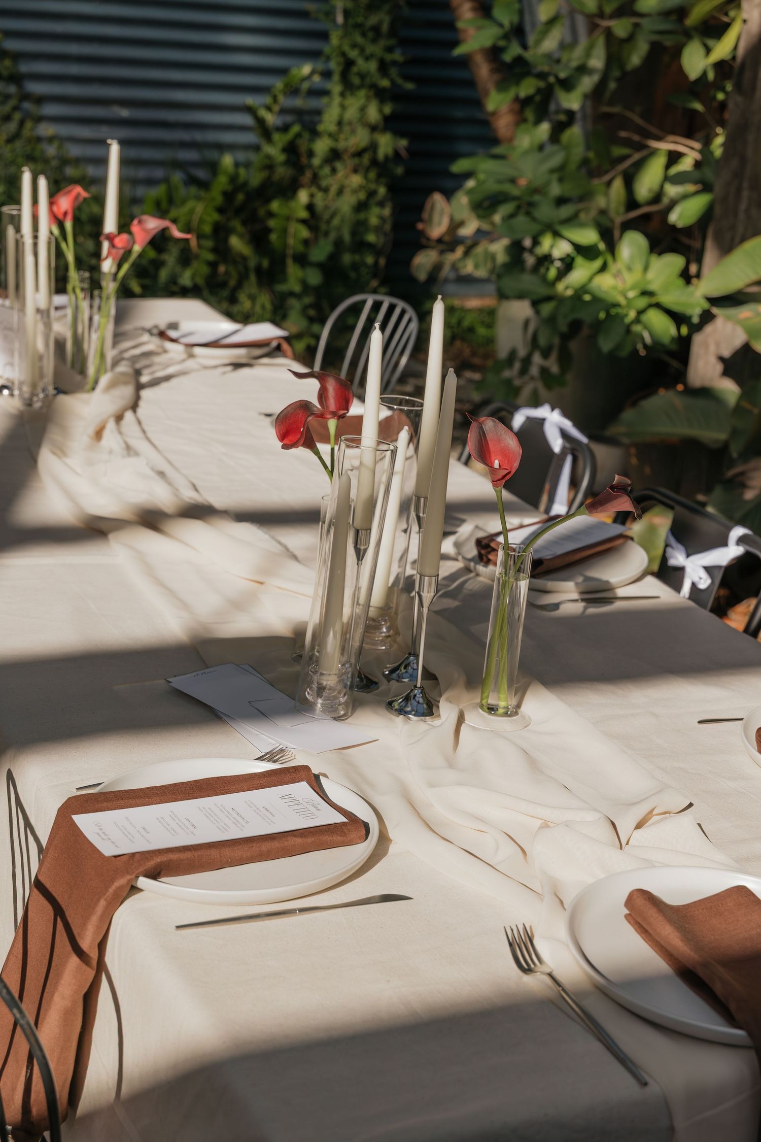 Elegant outdoor dining table set with white linen, terracotta napkins, candles, and red anthurium flowers in glass vases.