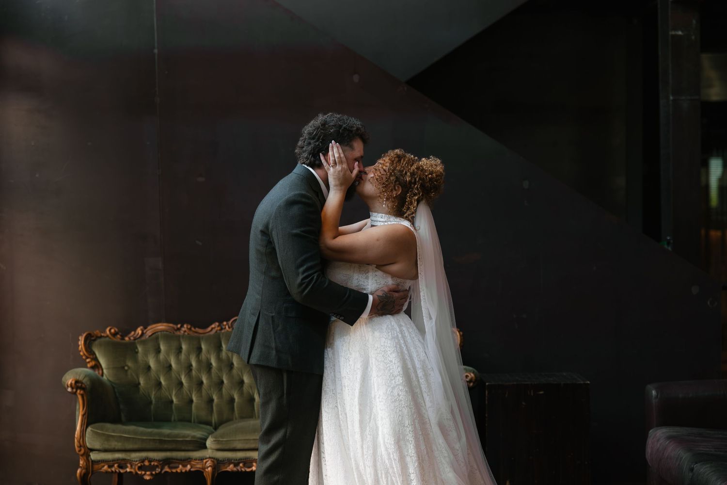 Bride and groom share a romantic first kiss in elegant wedding attire beside a vintage green velvet sofa.