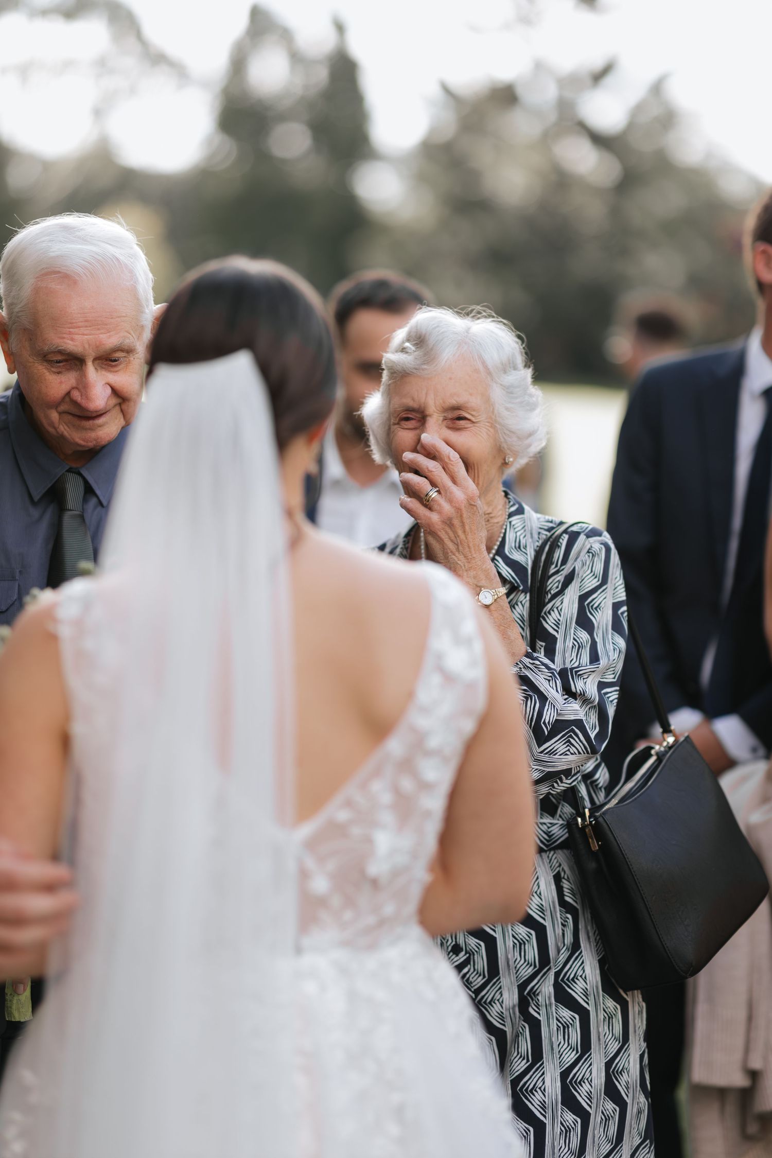 Elderly woman covers her mouth in emotional reaction seeing bride in lace wedding dress at outdoor wedding reception.