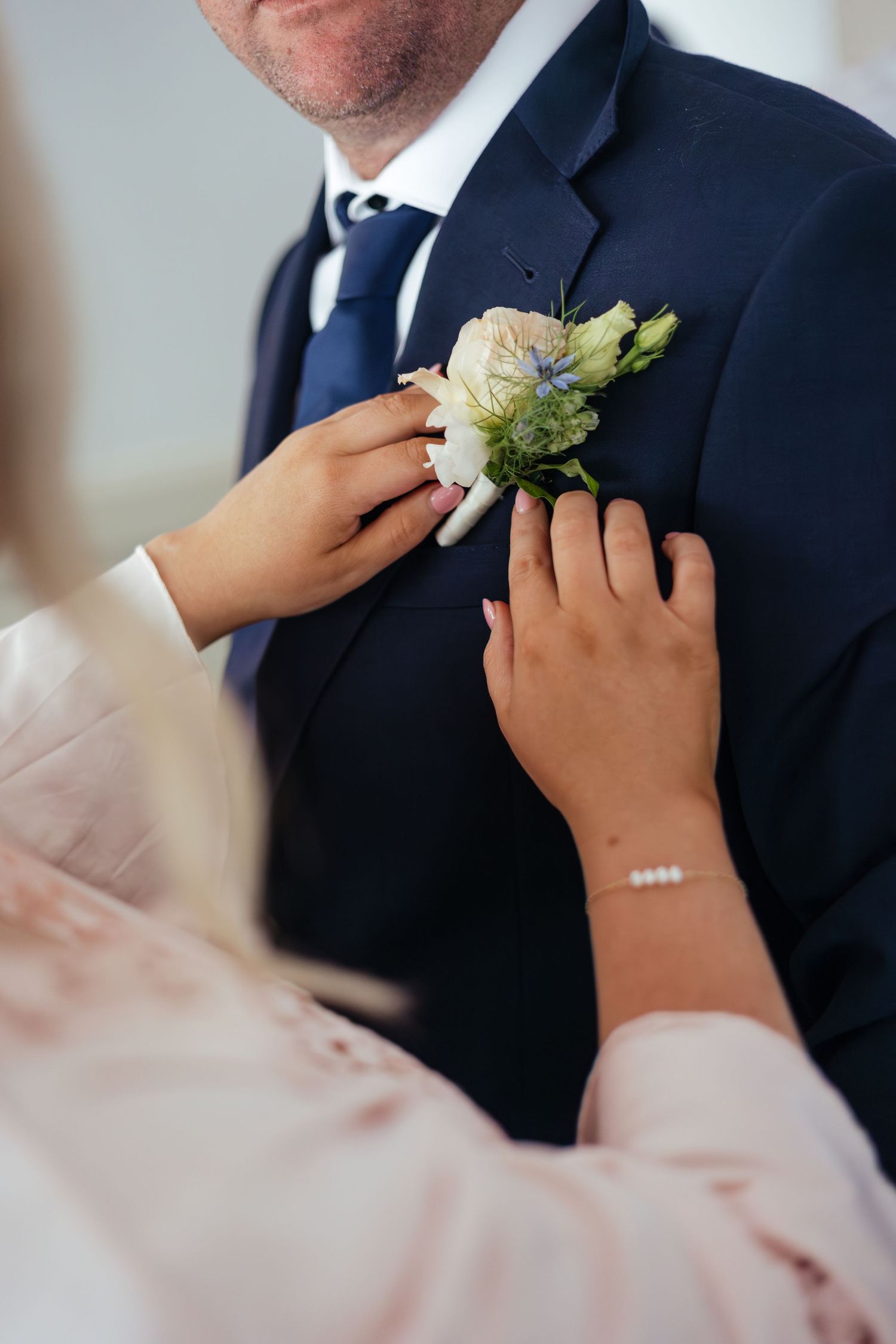 Hands pinning a white and blue floral boutonniere on a groom's navy blue suit lapel at a wedding.