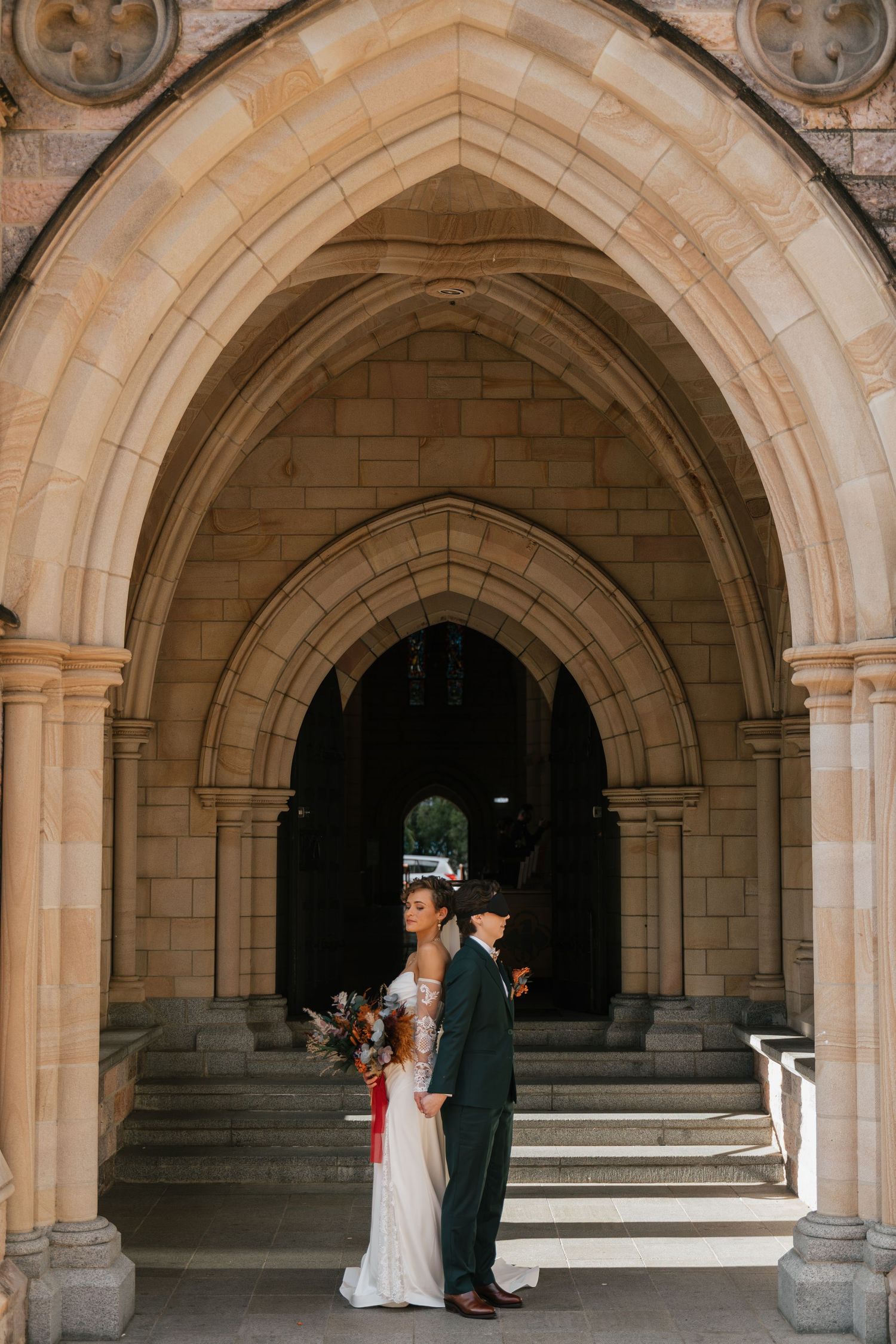 Bride and groom embracing under Gothic stone arches at a church wedding venue, bride holding a boho bouquet.