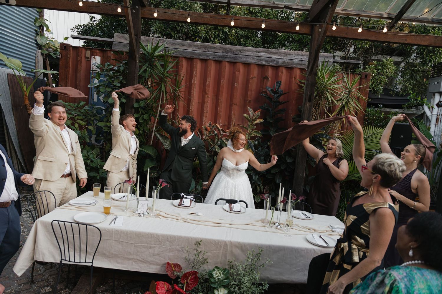 Wedding party celebrating at a decorated outdoor reception table with string lights and lush greenery backdrop.