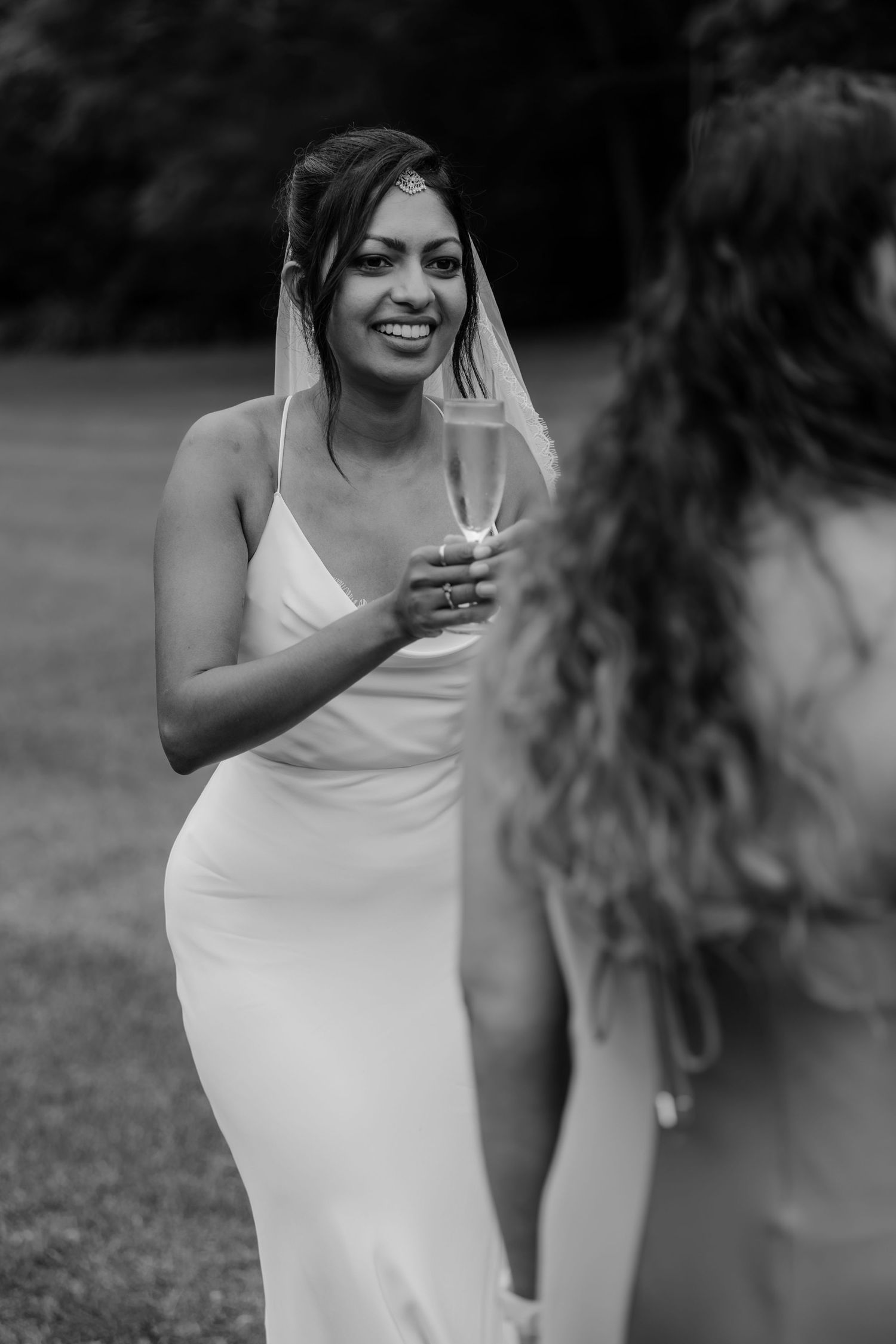 Black and white photo of a smiling bride in a white gown holding a champagne glass outdoors.